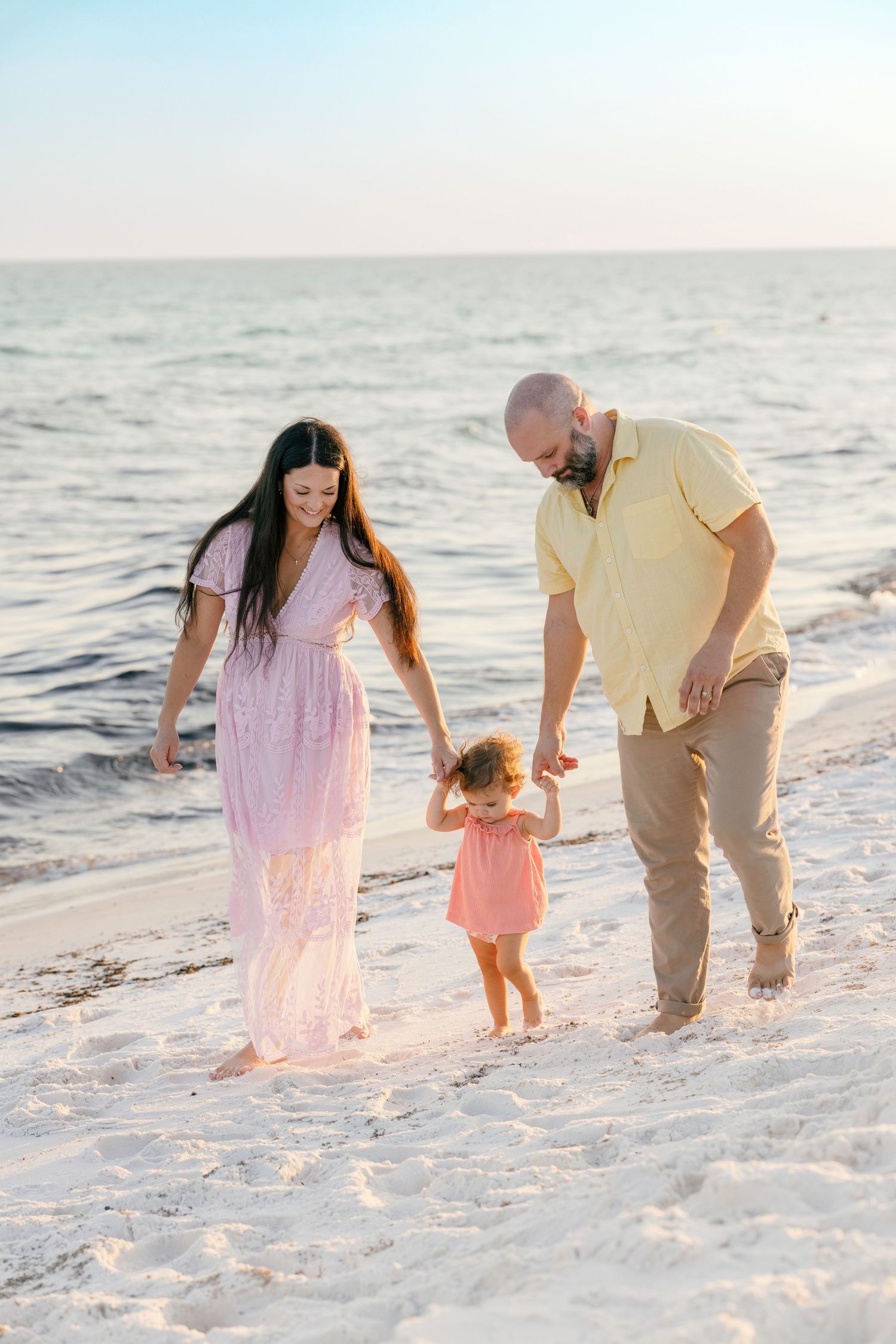 A happy family of three walks along a sandy beach at sunset, parents holding their toddler daughter's hands.