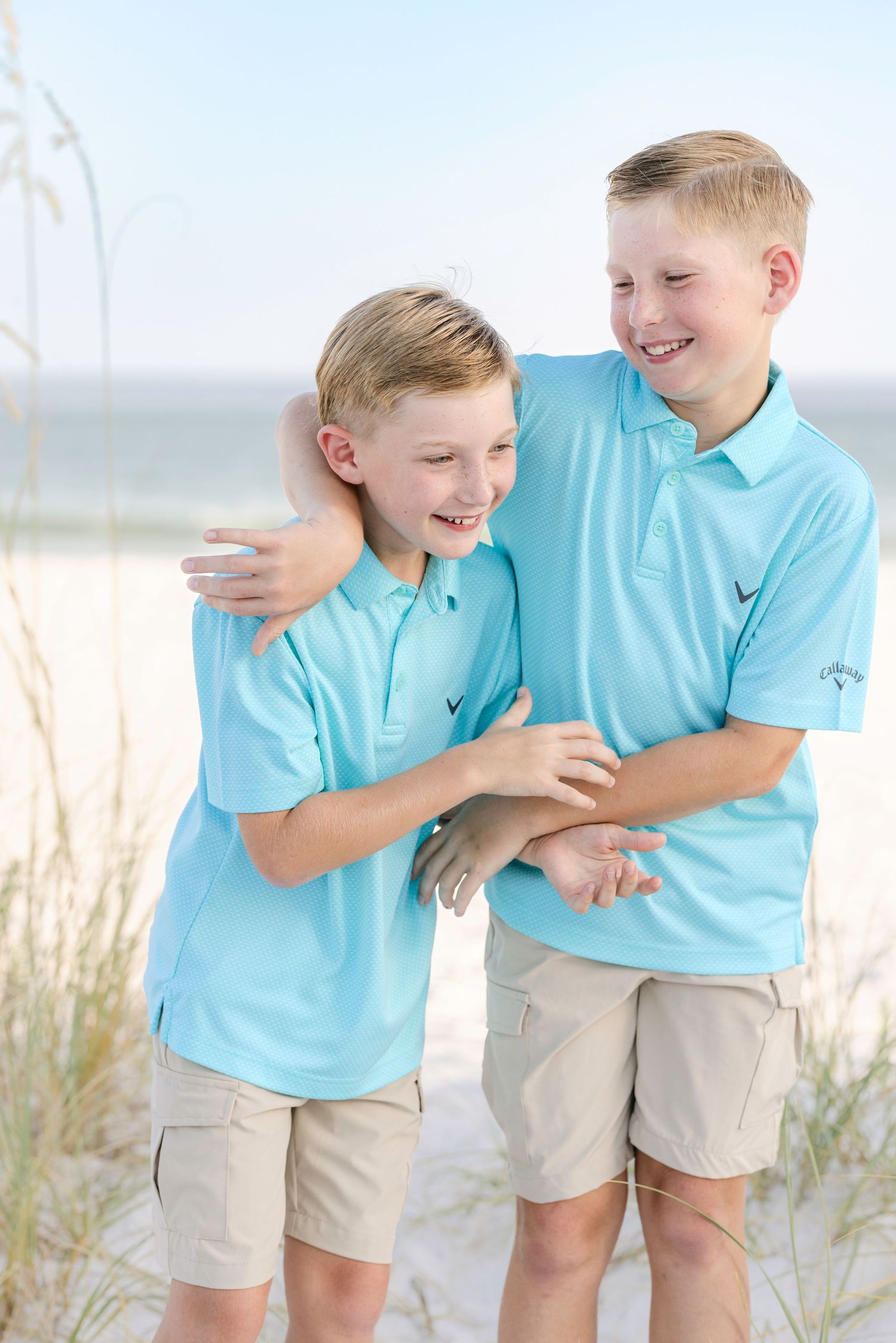 Two children in matching aqua polo shirts and khaki shorts share a playful moment on a sandy beach.