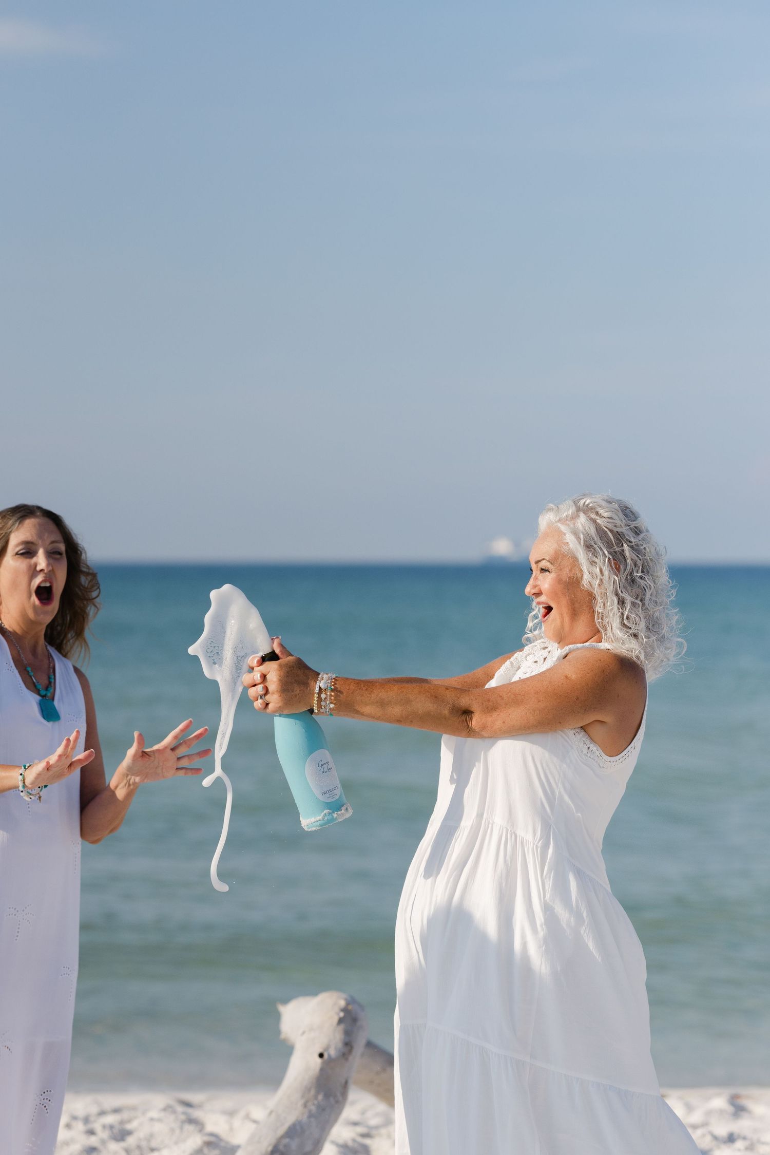 People in white dresses throw undergarments into the air while celebrating along a scenic beachfront.