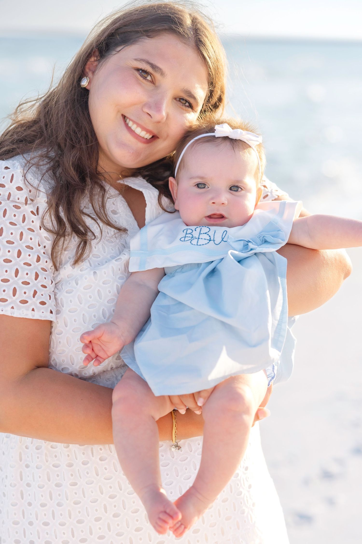 A woman in white lace holds a baby in a blue outfit during a beach photoshoot at sunset.
