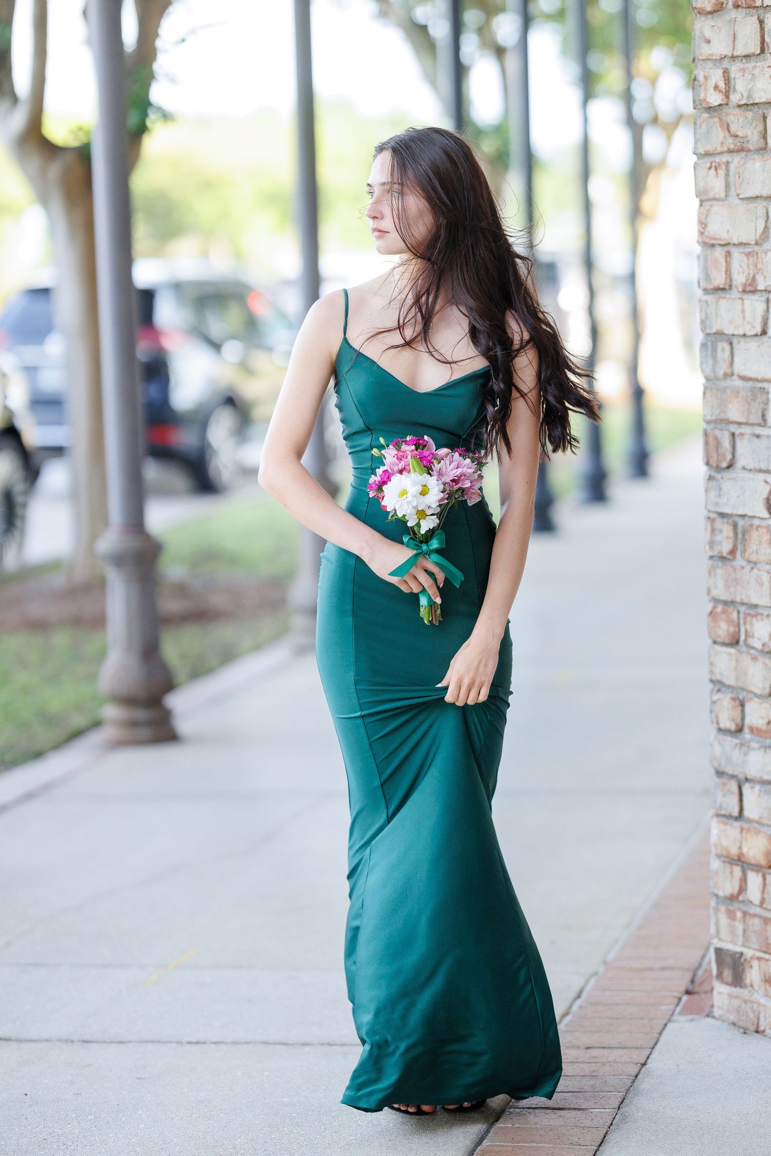 A woman in an elegant emerald green evening gown holds a pink floral bouquet while standing on a sidewalk.