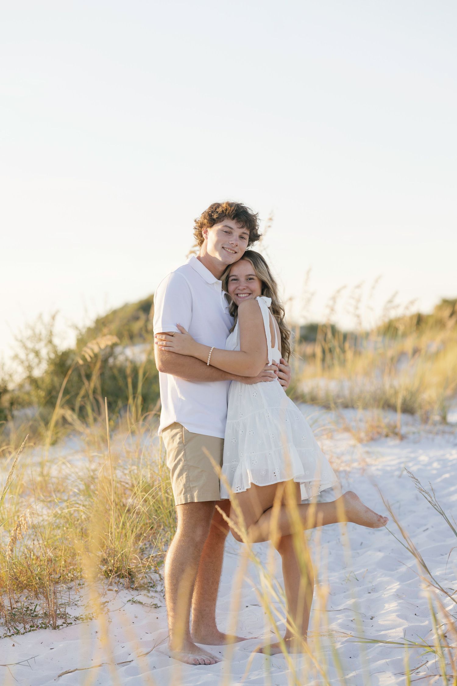 A couple embraces and laughs together on a sandy beach with grass and golden hour lighting in a romantic moment.