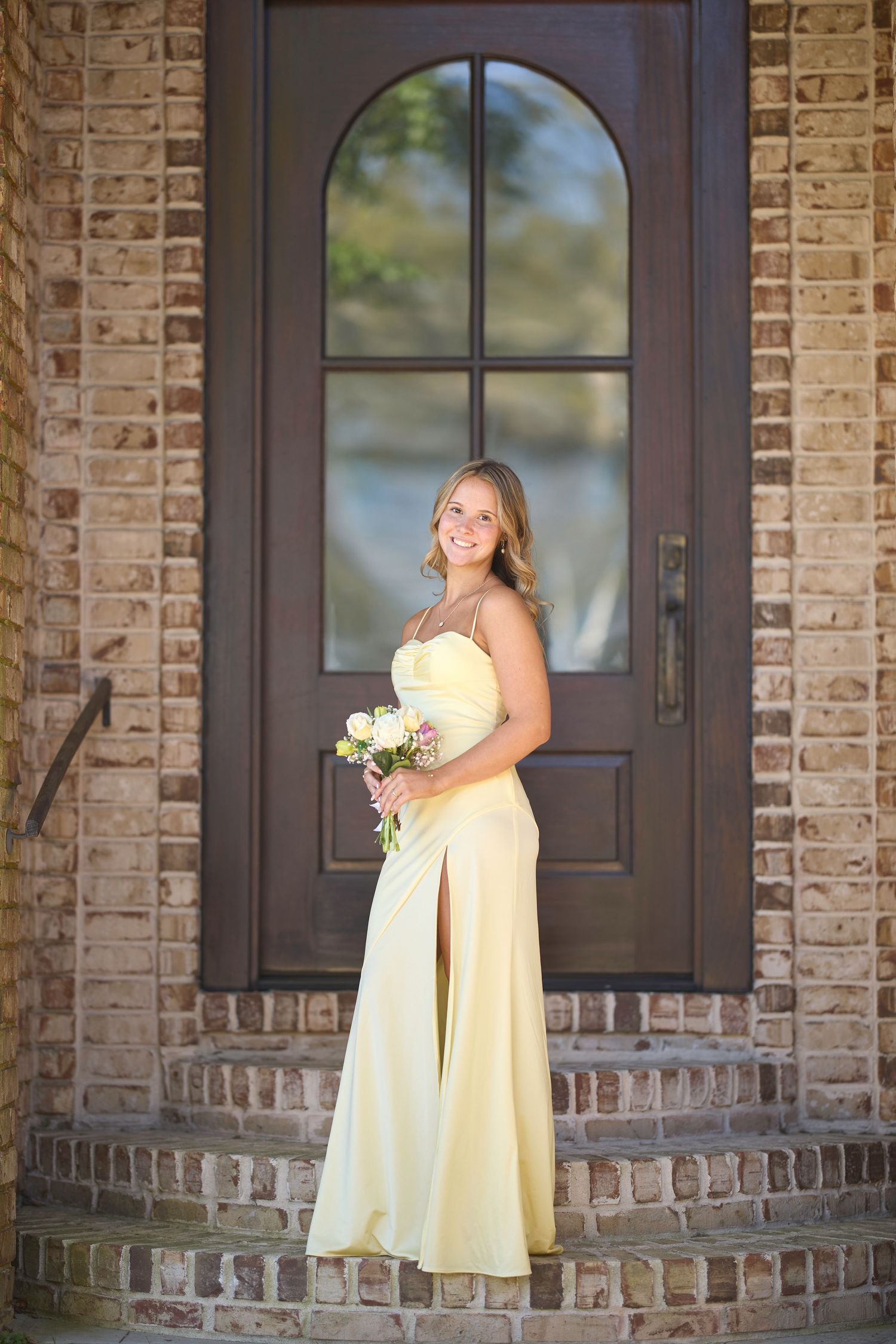 Figure in a pale yellow evening gown stands on brick steps holding a bouquet in front of an arched doorway.