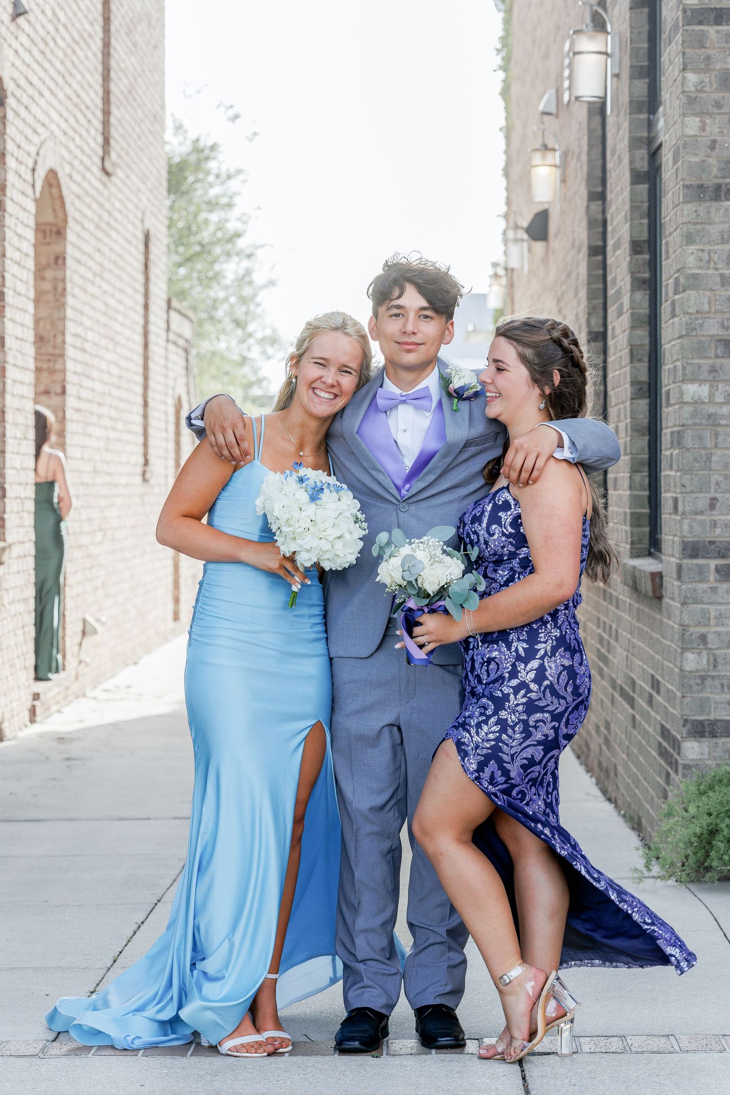Three people in formal attire pose together on a sidewalk, wearing blue and navy dresses and a gray suit with bouquets.