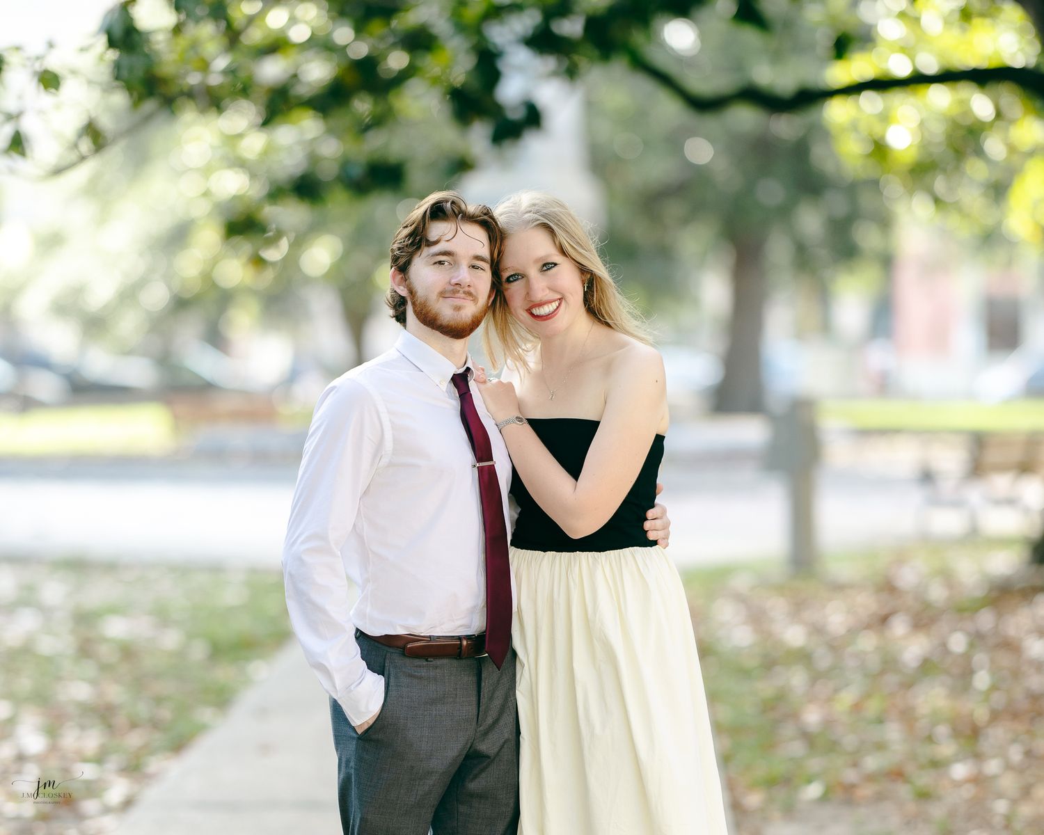 A couple embraces in an outdoor setting with fall foliage visible in the background.