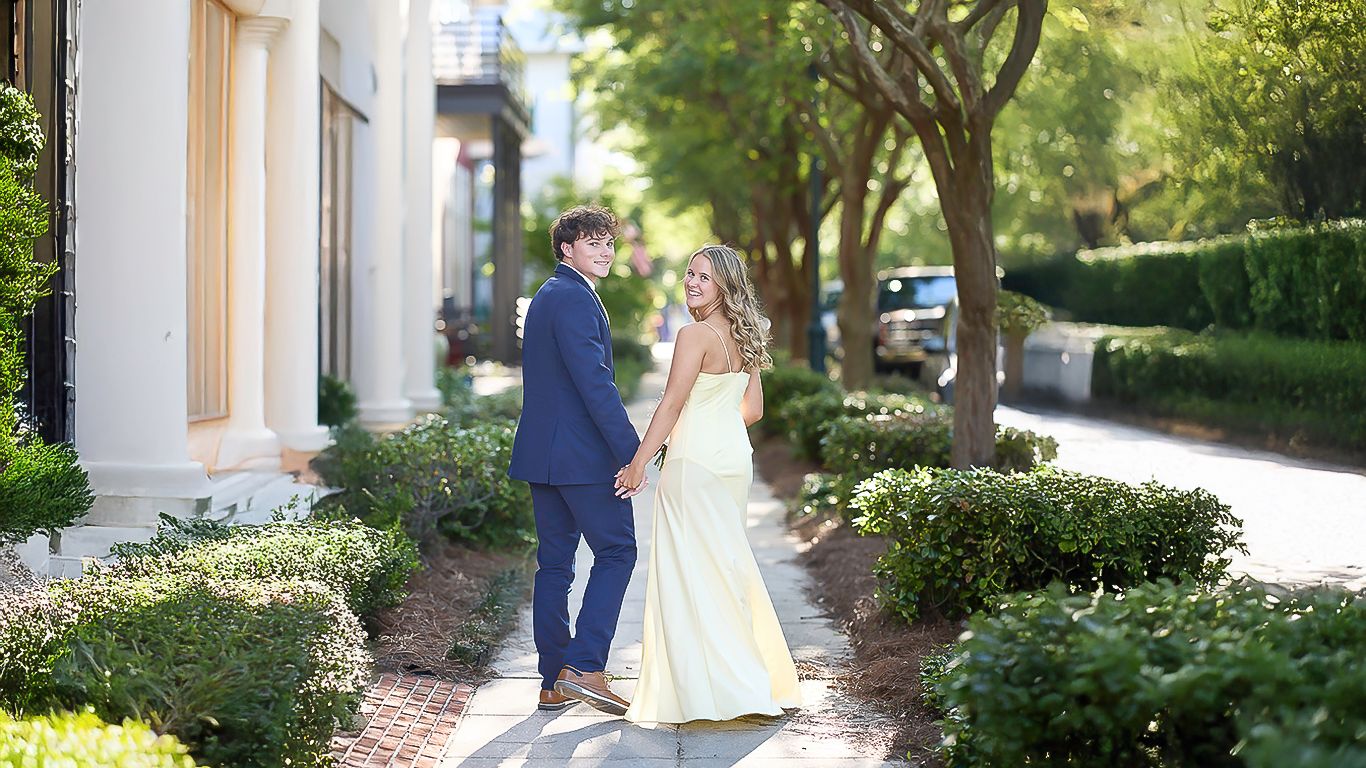 Couple in wedding attire walking along garden path at elegant venue on sunny day.
