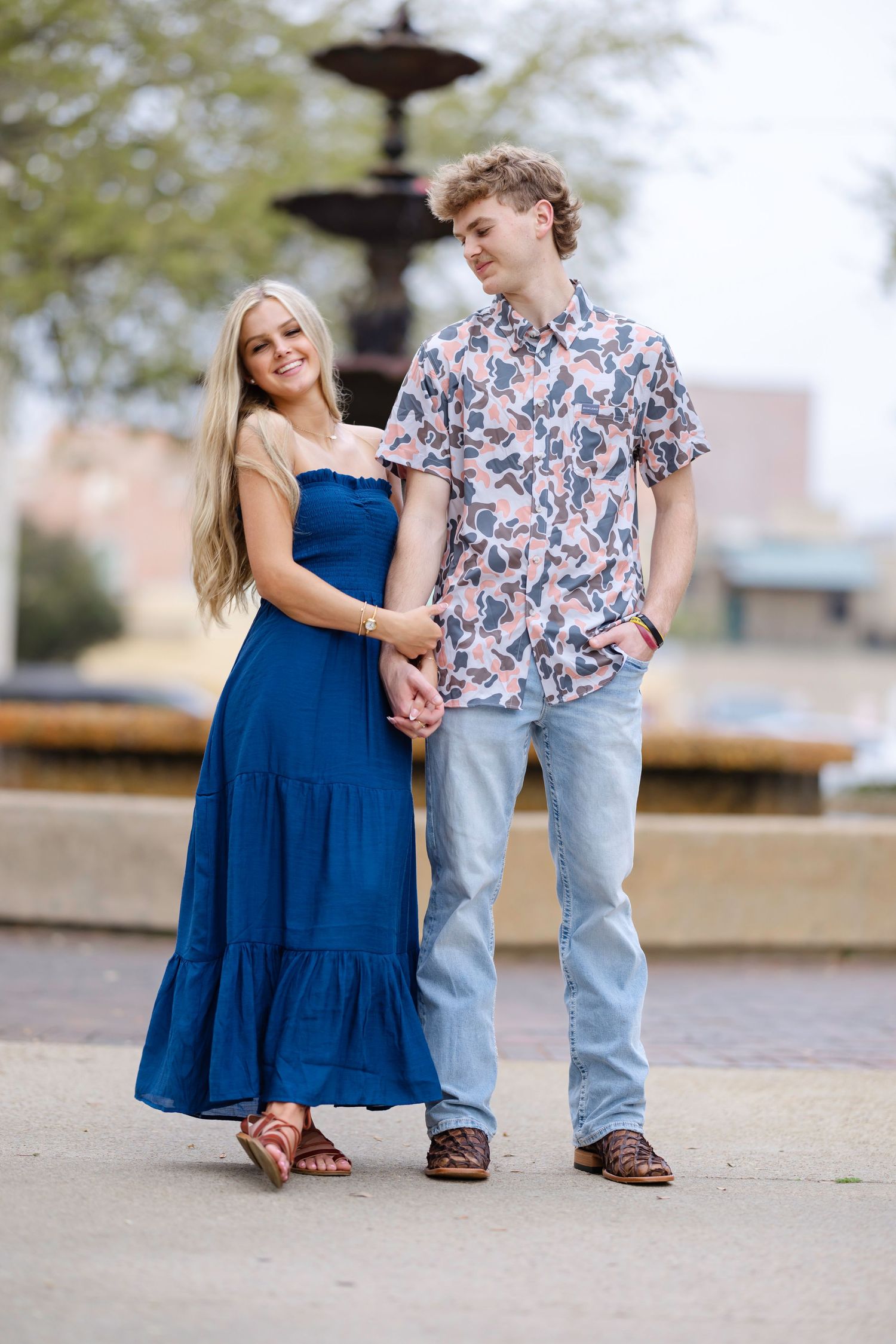 A couple in blue clothing poses playfully together on a paved outdoor area near a fountain.