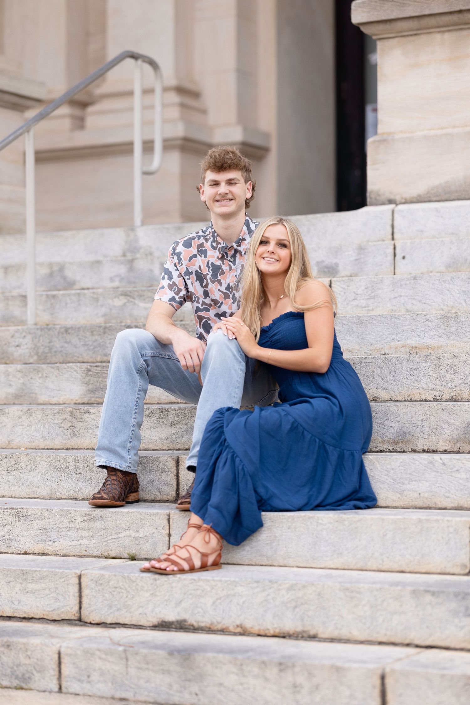 A couple in casual attire poses together on stone steps outside a grand building.
