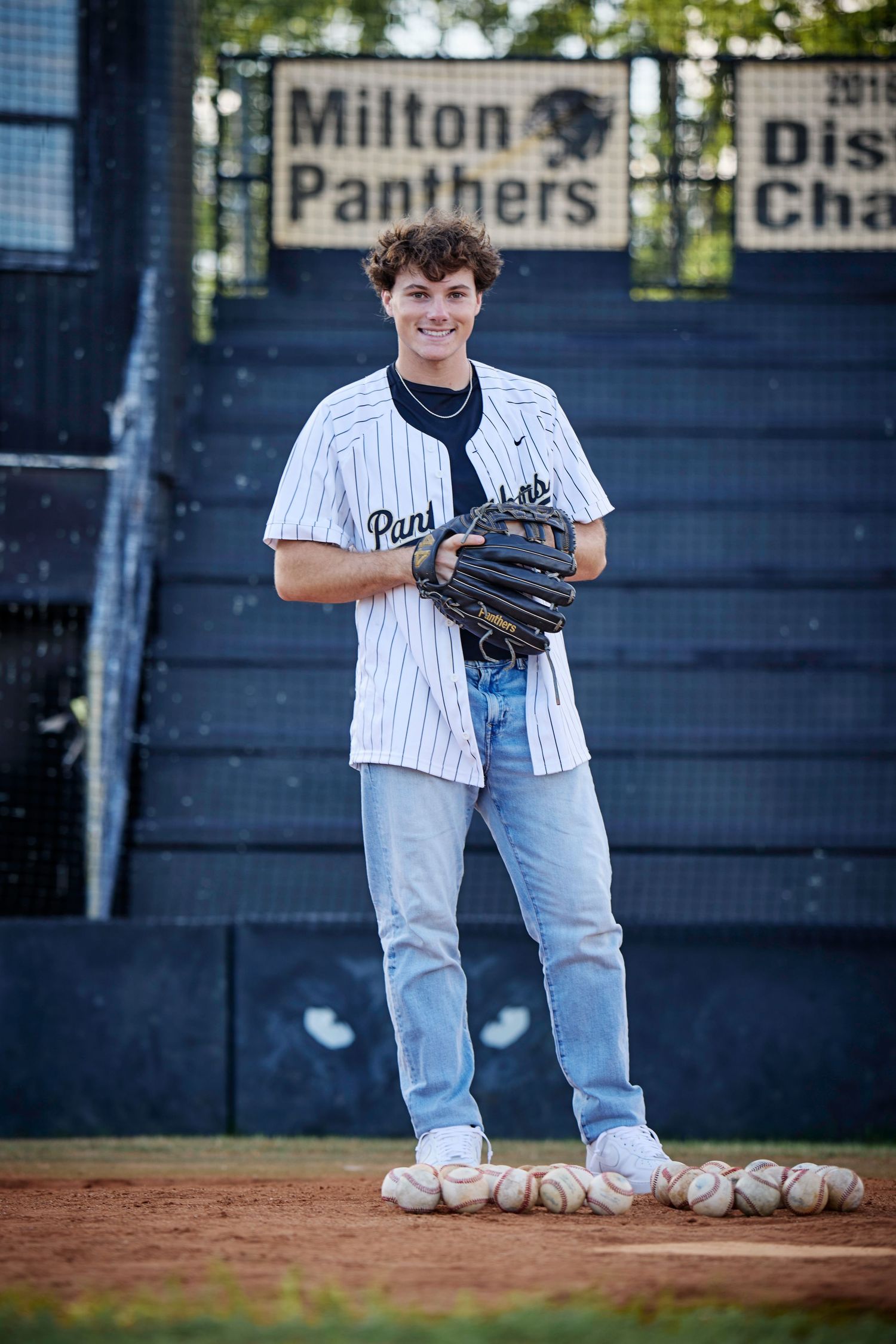 Baseball player stands by Milton Panthers sign holding glove with baseballs at feet.