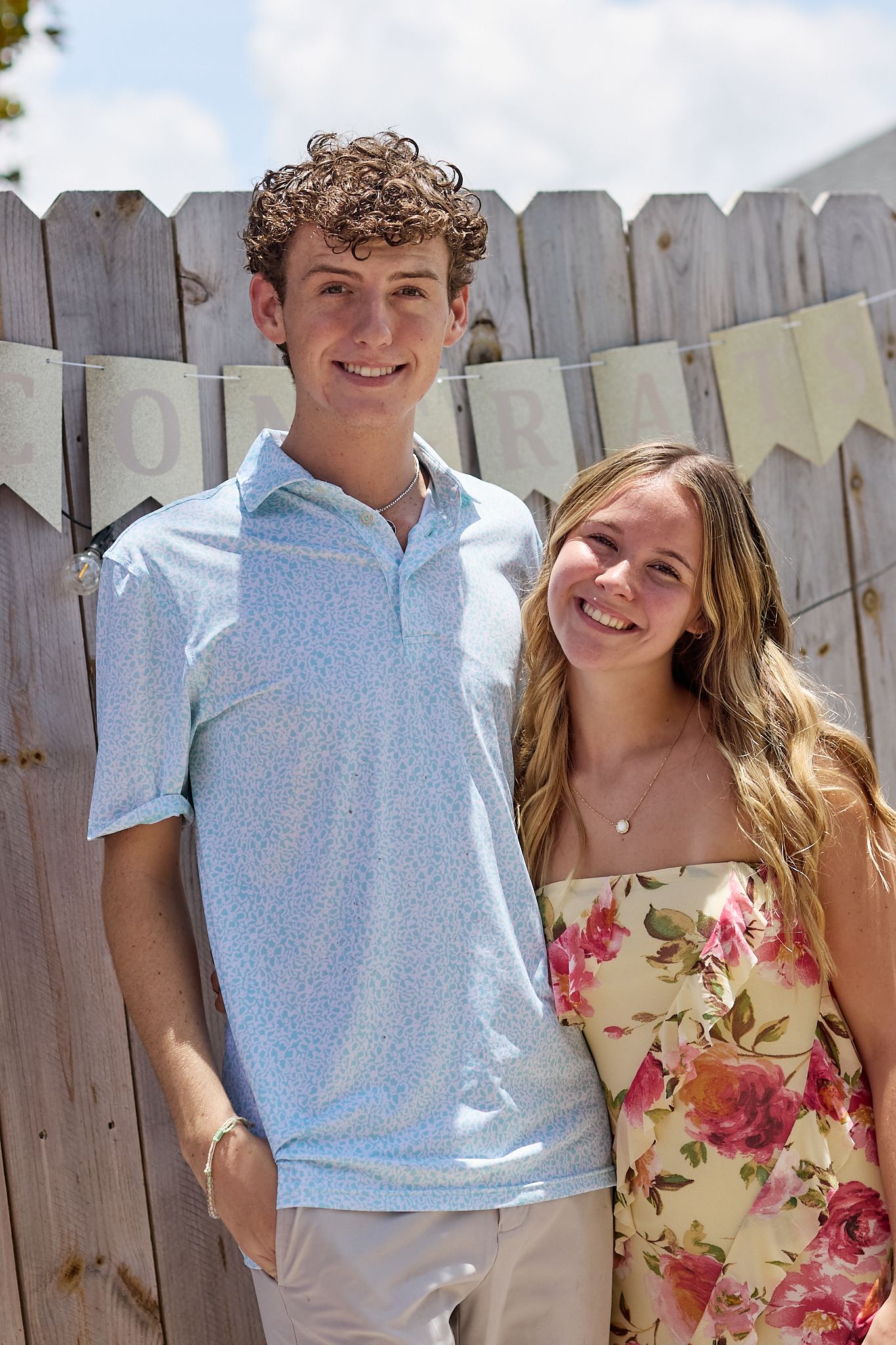 A couple poses outdoors in front of a wooden fence on a sunny day.