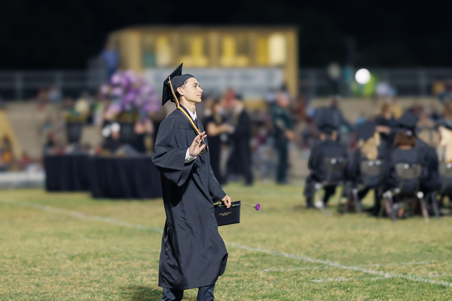 Graduate in black cap and gown walks across outdoor stadium field during evening ceremony.