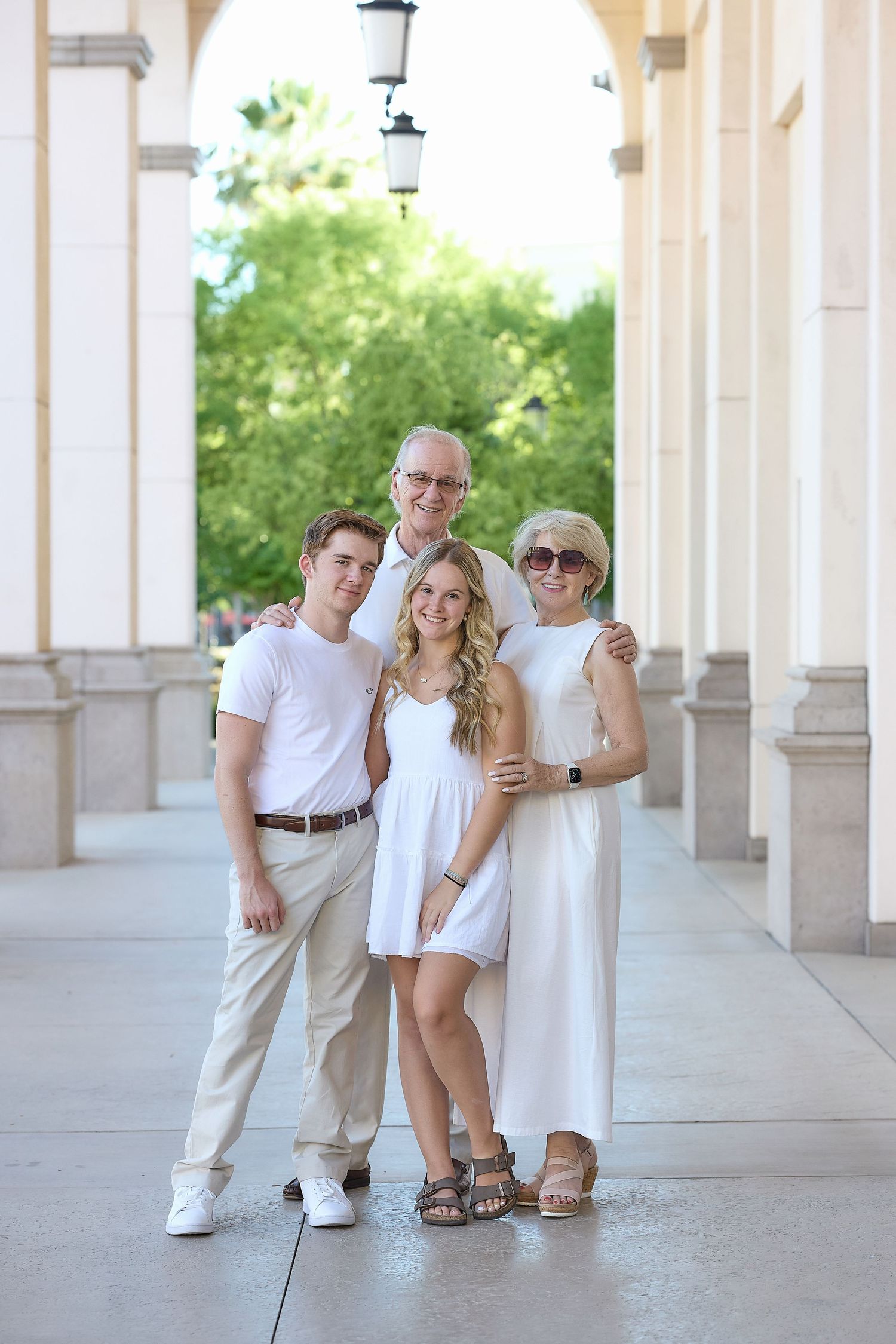 A family wearing coordinated white and beige outfits poses together under an elegant columned archway.