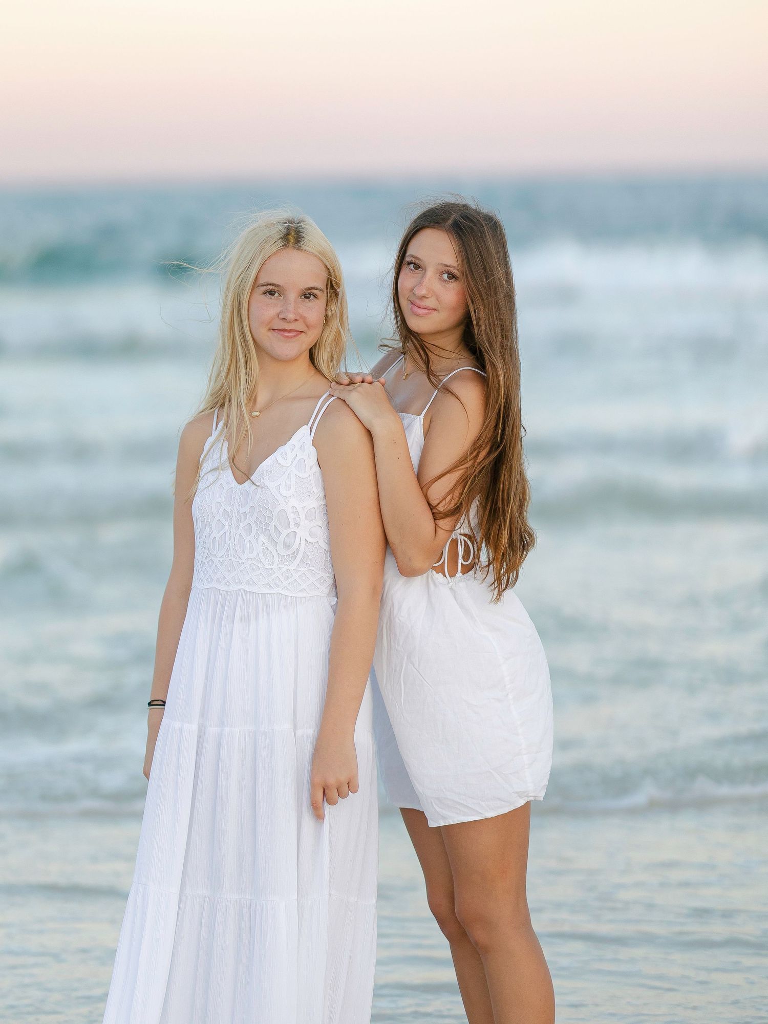 Two friends in flowing white dresses pose together on a sandy beach with turquoise waves behind them.