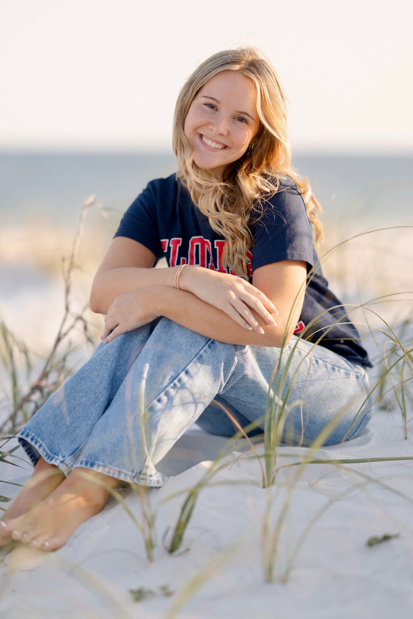 Smiling blonde teen girl sitting on white sand beach dunes wearing jeans and navy college shirt on sunny day.
