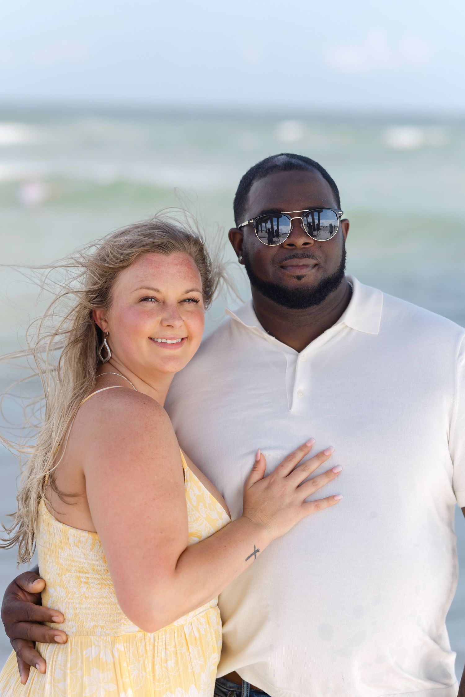 Interracial couple embracing on a sunny beach, woman in yellow dress with blonde hair, man in white shirt wearing sunglasses.
