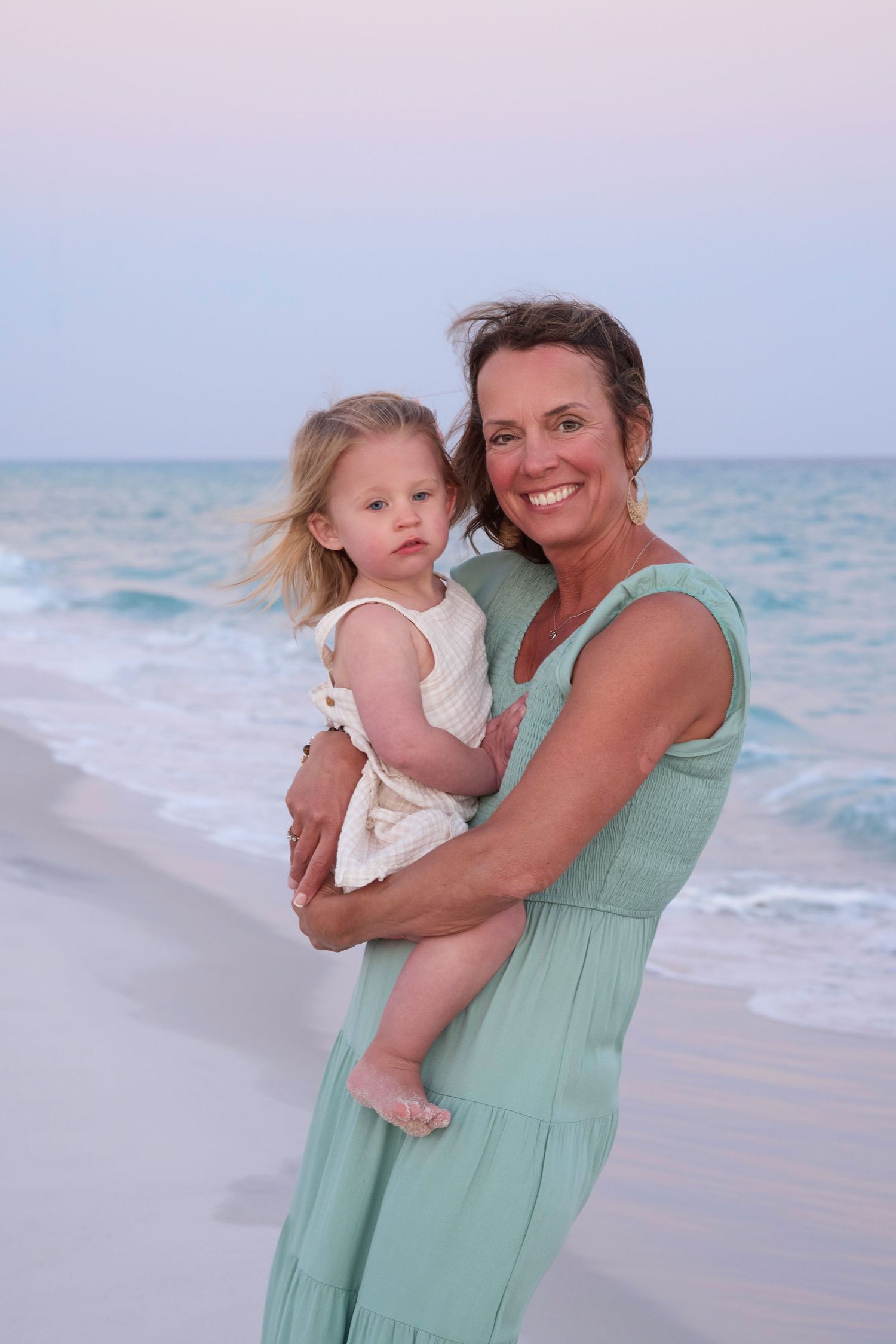 A smiling woman in a teal dress holds a young blonde girl on a sandy beach at sunset with ocean waves behind them.