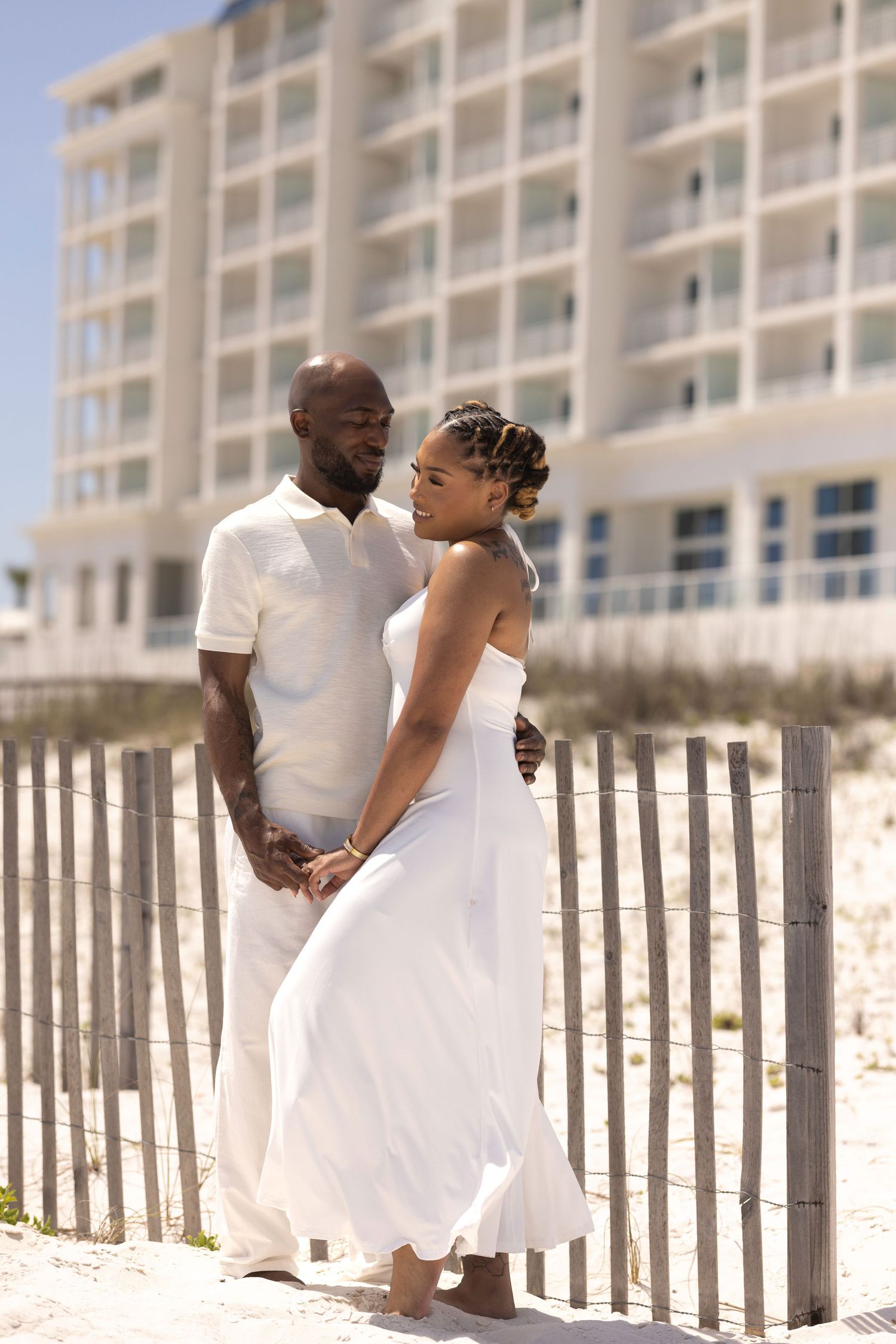 African American couple in white outfits holding hands on sandy beach with coastal building backdrop.