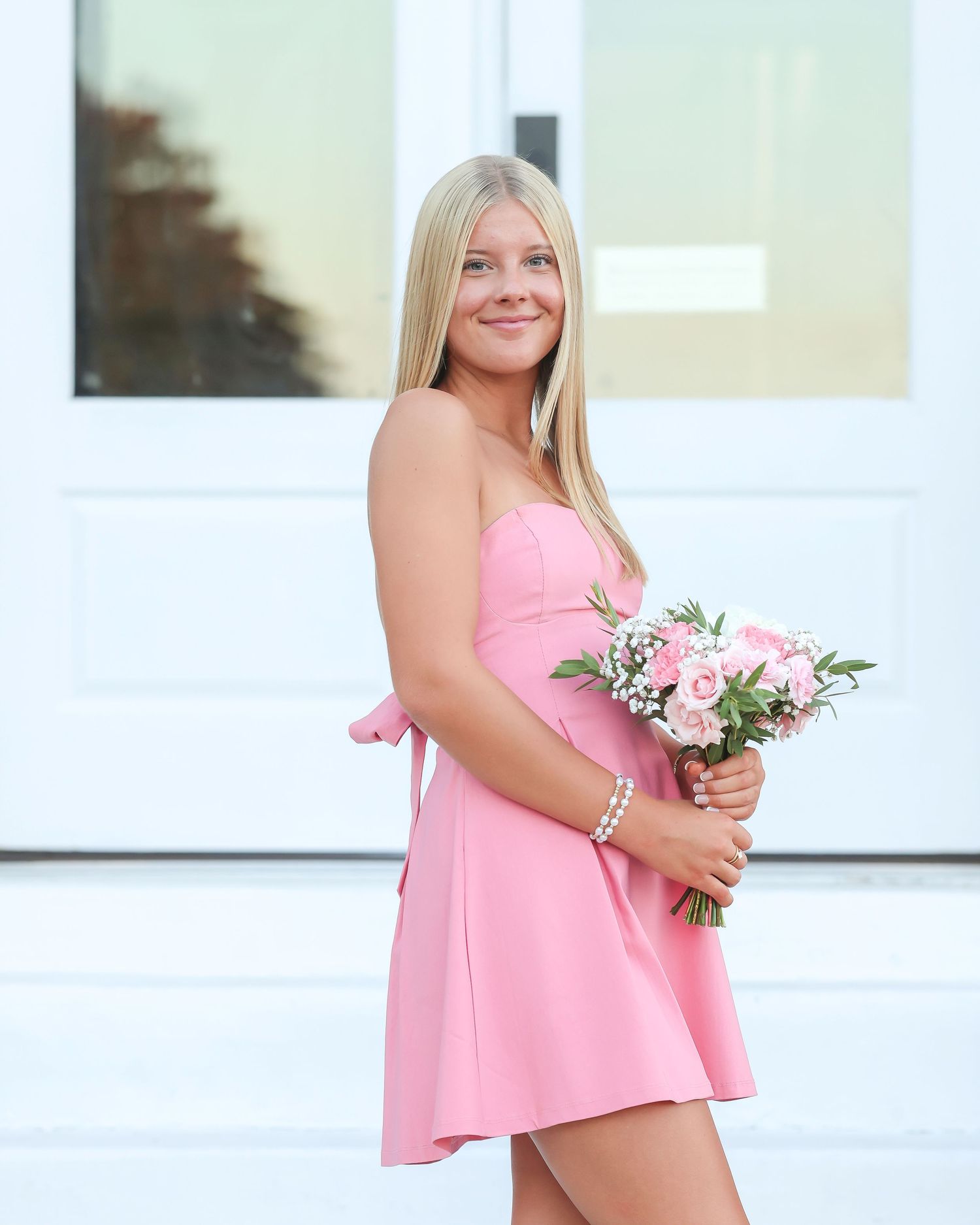 A person in a pink strapless dress holds a small bouquet of white and pink flowers next to a white wall.