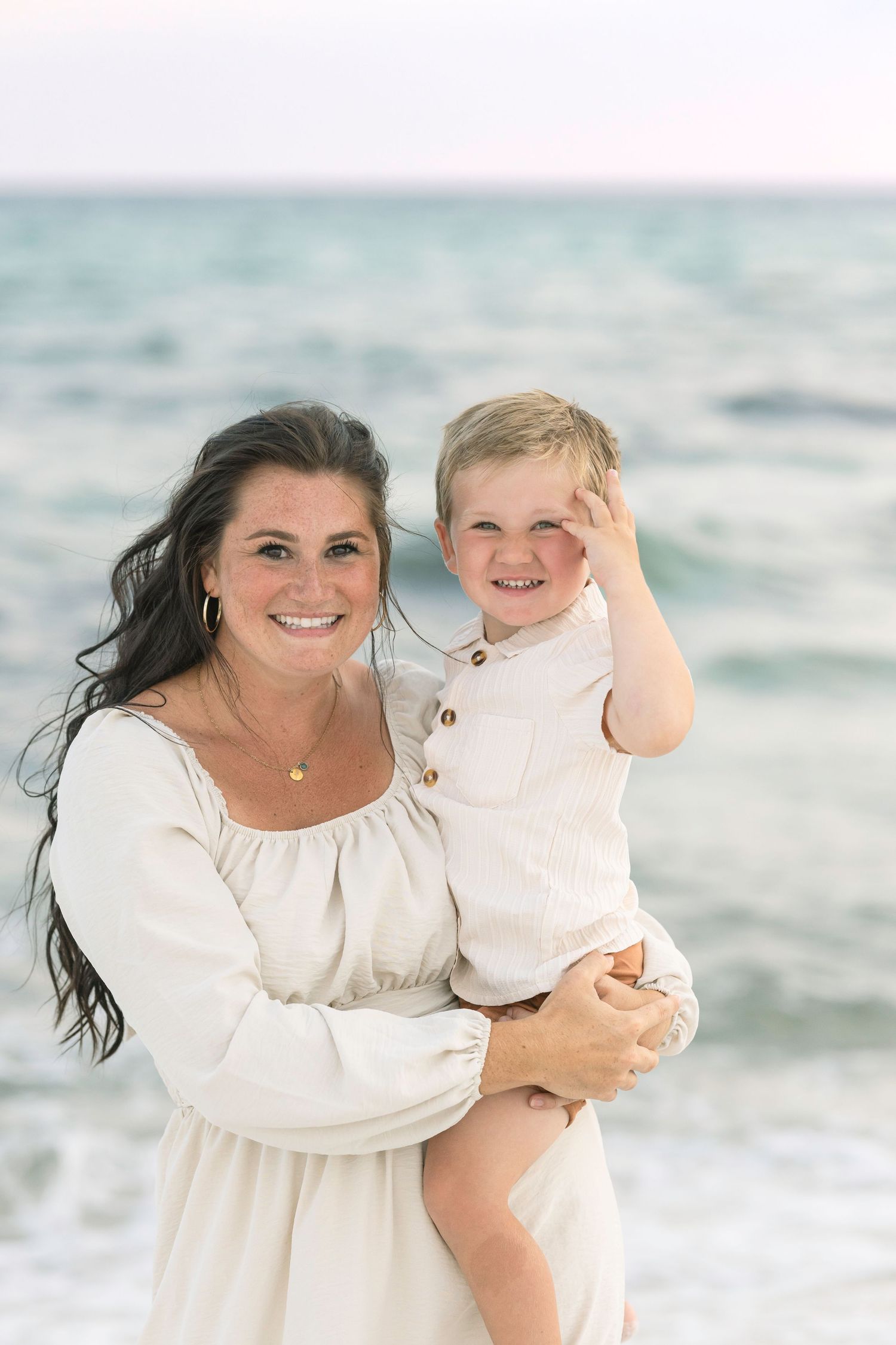 A smiling woman in a white dress holds a young blond boy at the beach, both looking happy with ocean waves behind them.