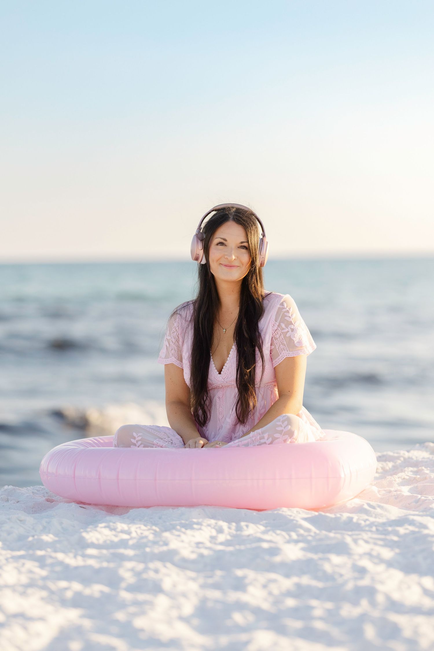 Woman in pink lace dress sitting on white sand beach with pink inflatable ring and headphones.