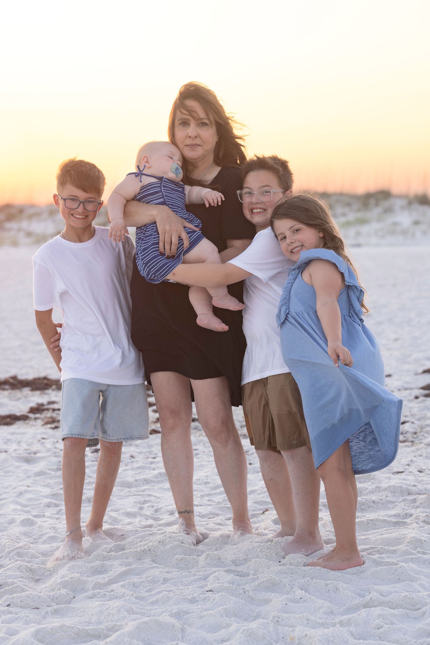 A mother holds a baby while four children surround her on a beach at a warm sunset.