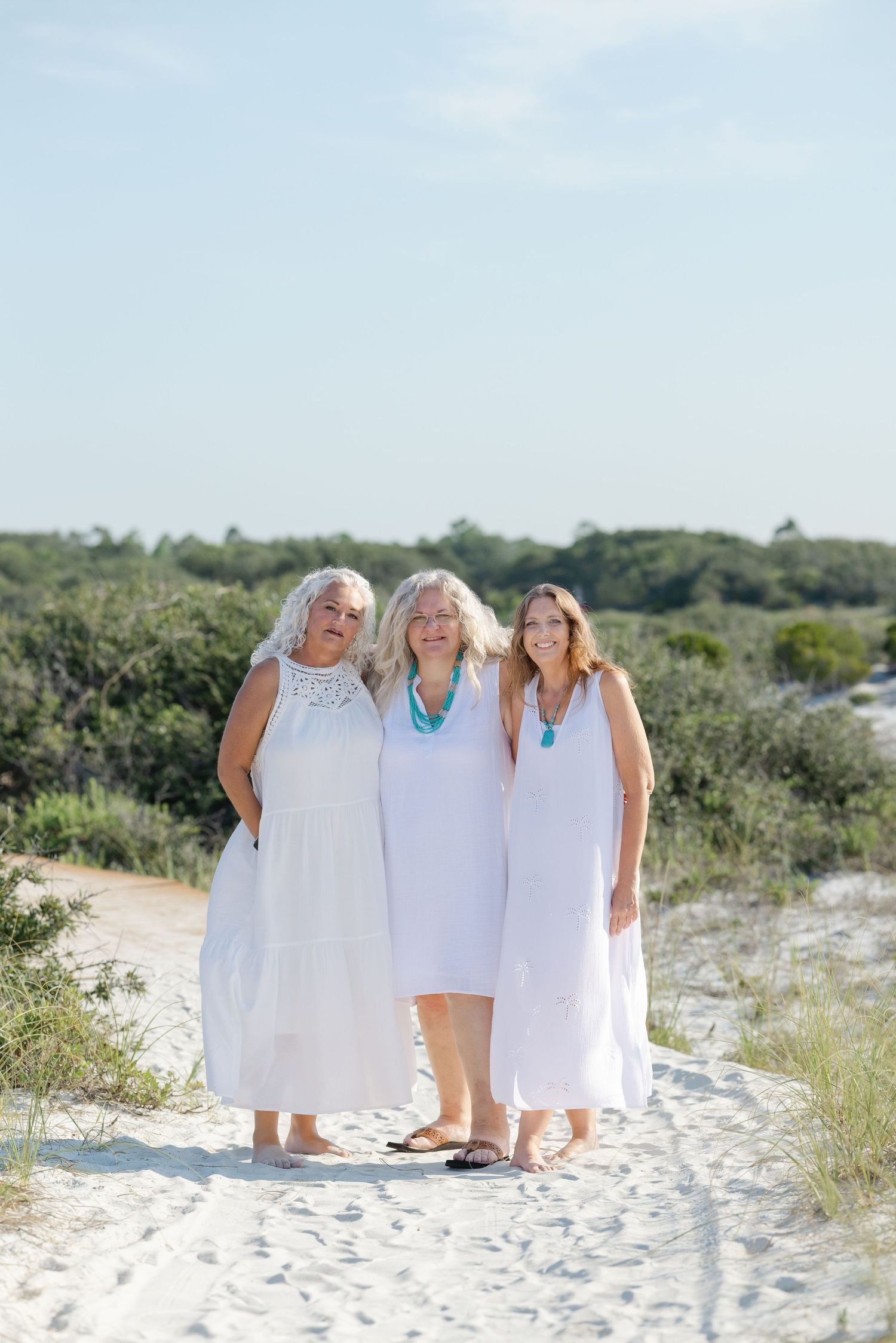 Three people wearing white dresses pose together on a sandy dune with green coastal vegetation in the background.