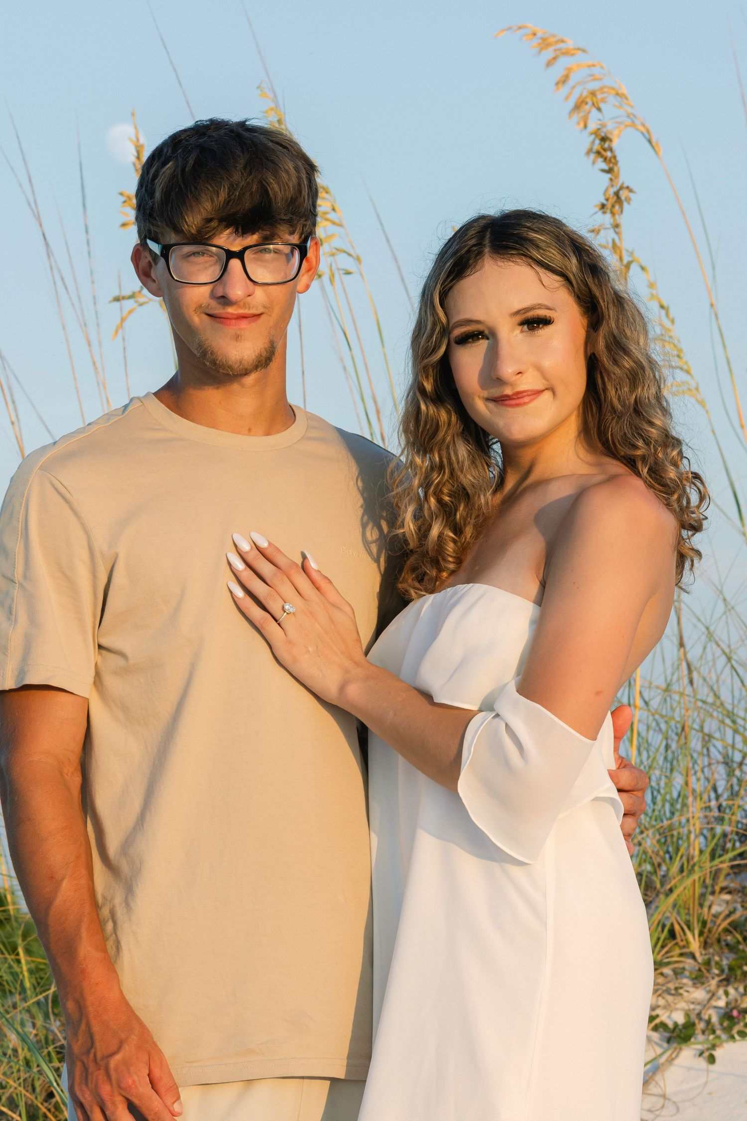 A couple in casual beachwear poses together against coastal grass and blue sky.