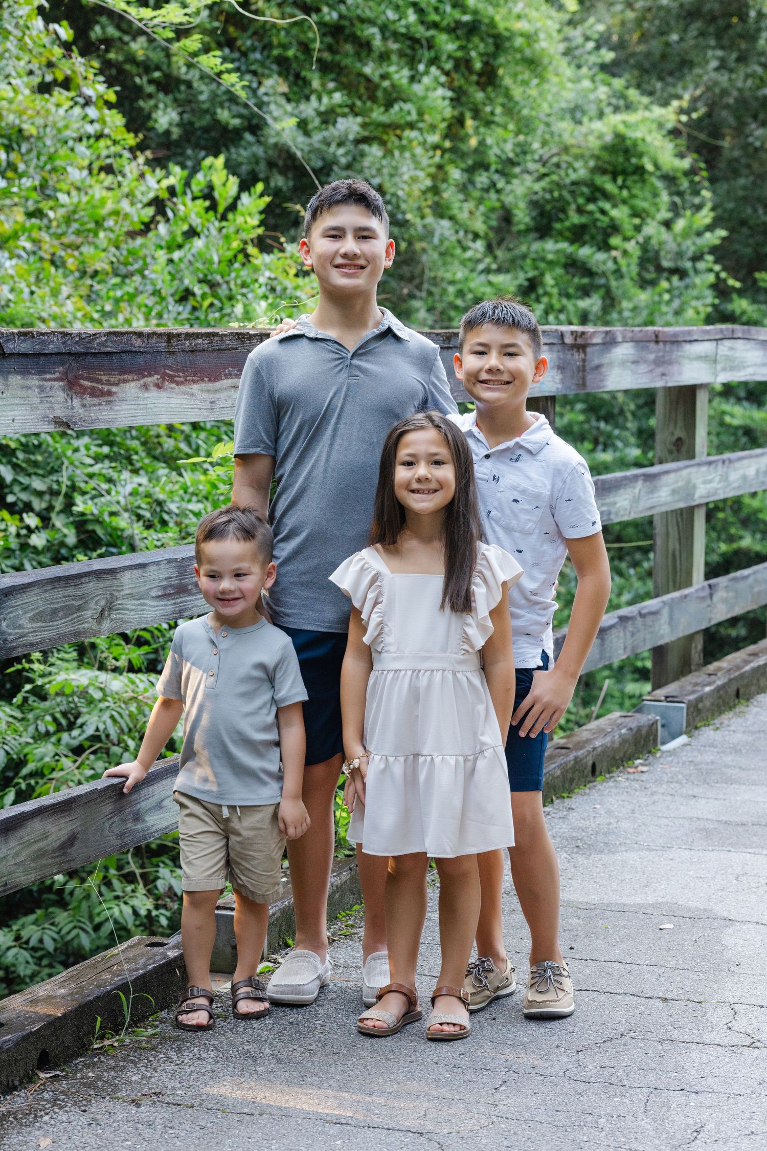 Siblings stand together on a wooden bridge walkway surrounded by lush green foliage on a sunny day.