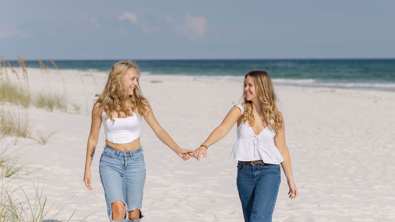Two people in jeans and white tops holding hands while walking together on a sunny sandy beach.