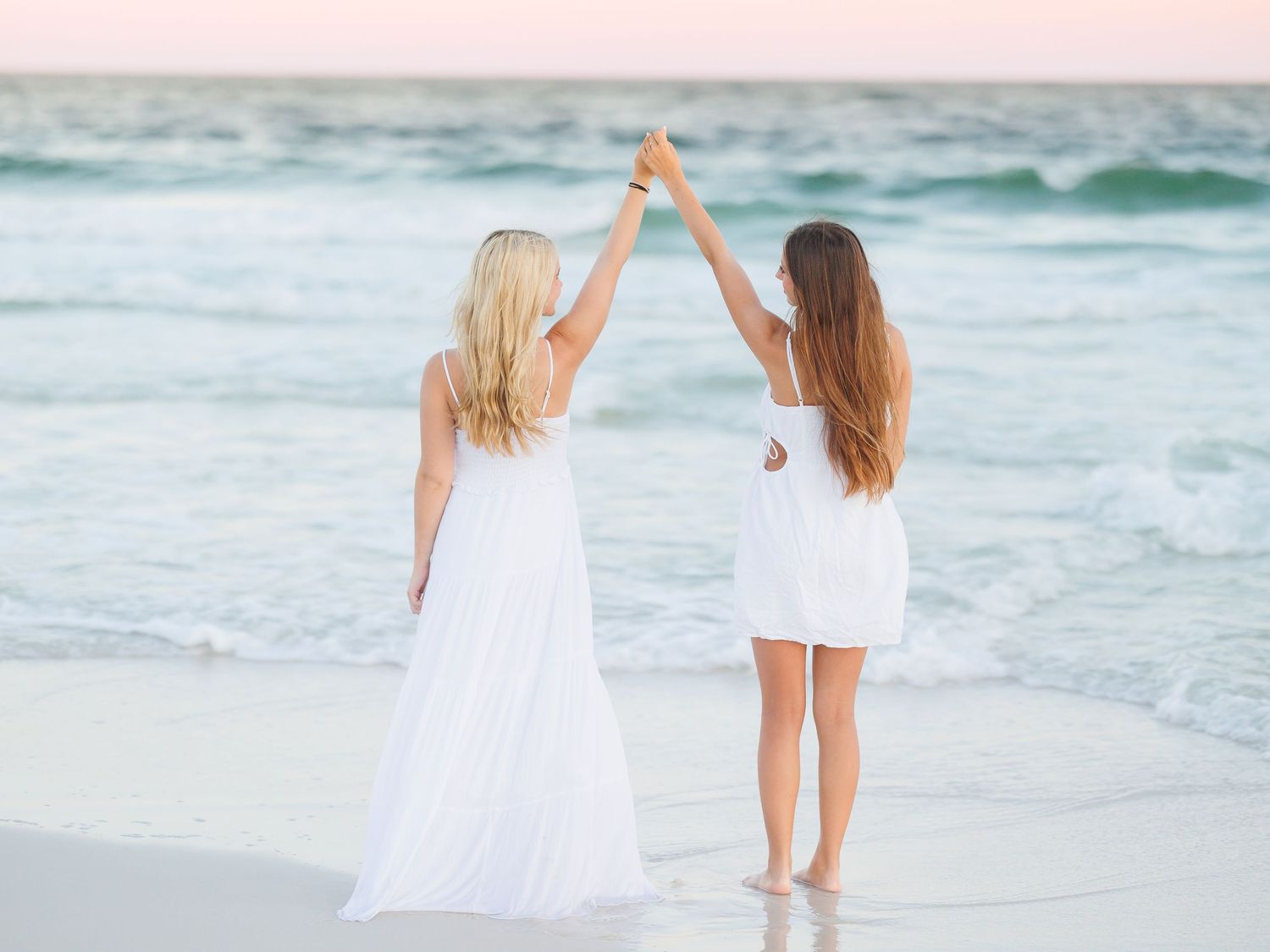 Two people in white dresses hold hands up while standing on beach at sunset.