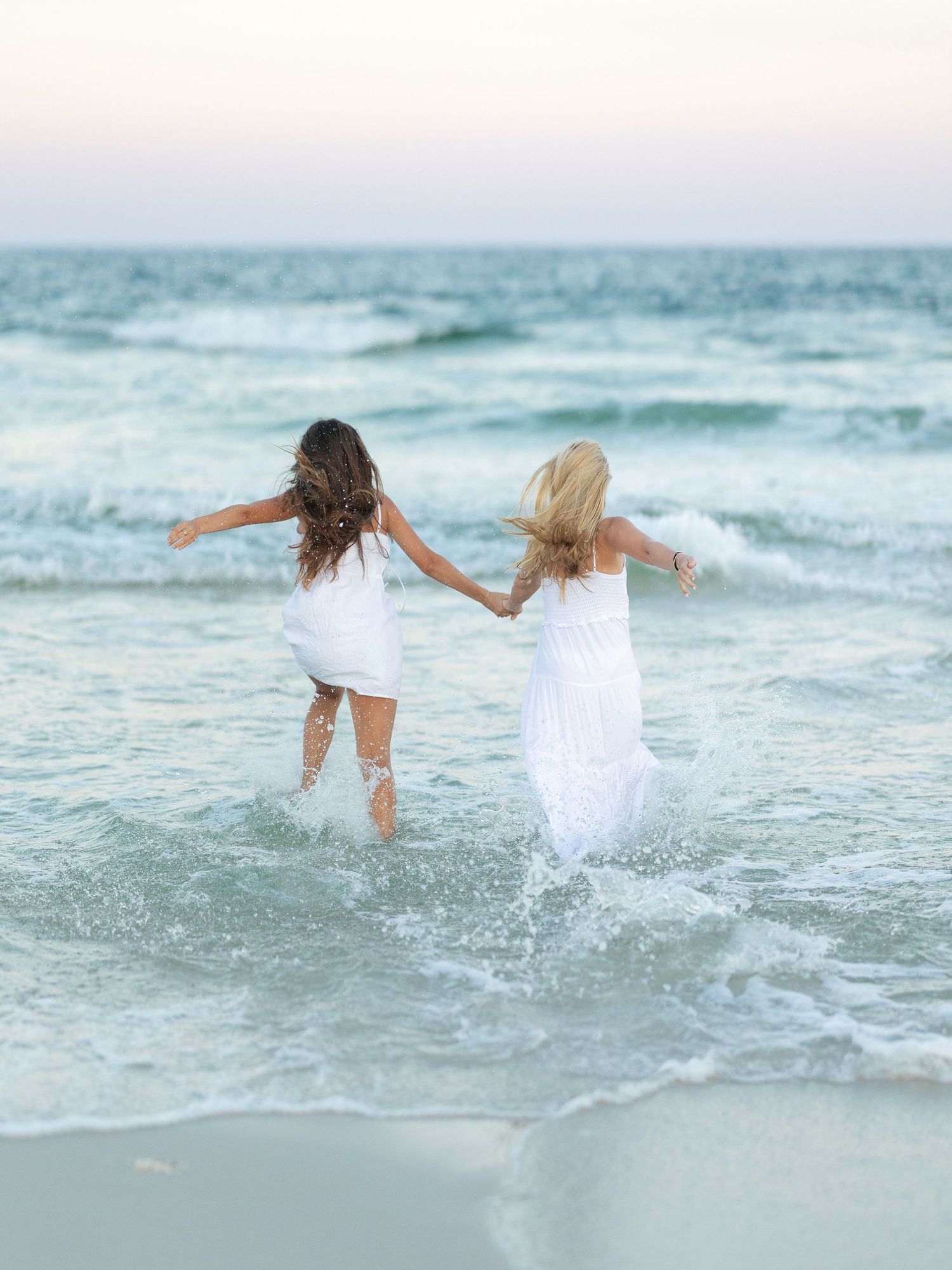 Two friends splashing through ocean waves while wearing white dresses.
