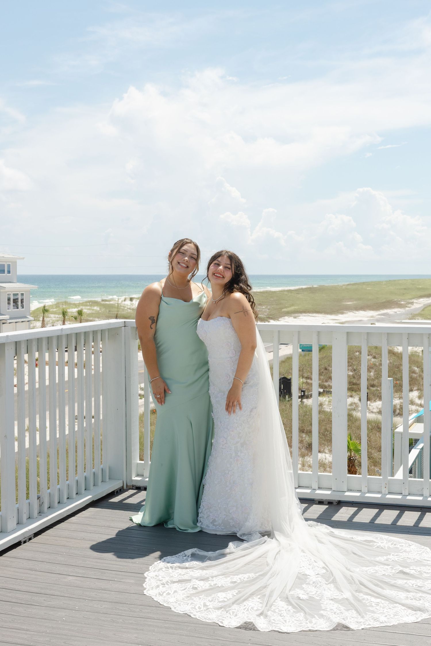 A seaside wedding photo featuring a bride and bridesmaid on a beach house balcony.