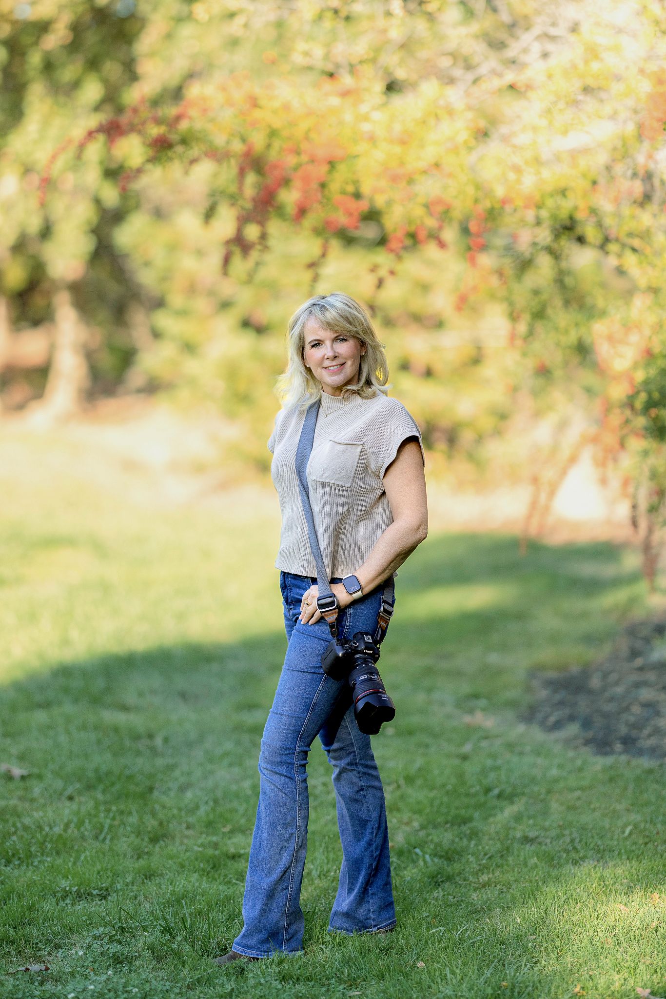Person in jeans and sleeveless top poses playfully on grassy hillside with autumn foliage in background.