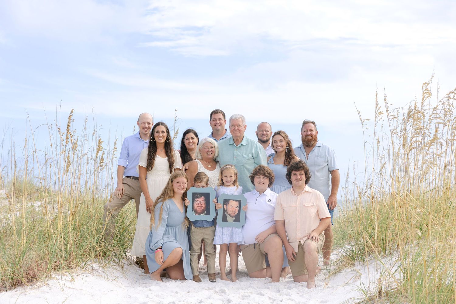 Extended family poses together on a sunny beach with sea oats and sand dunes in the background.
