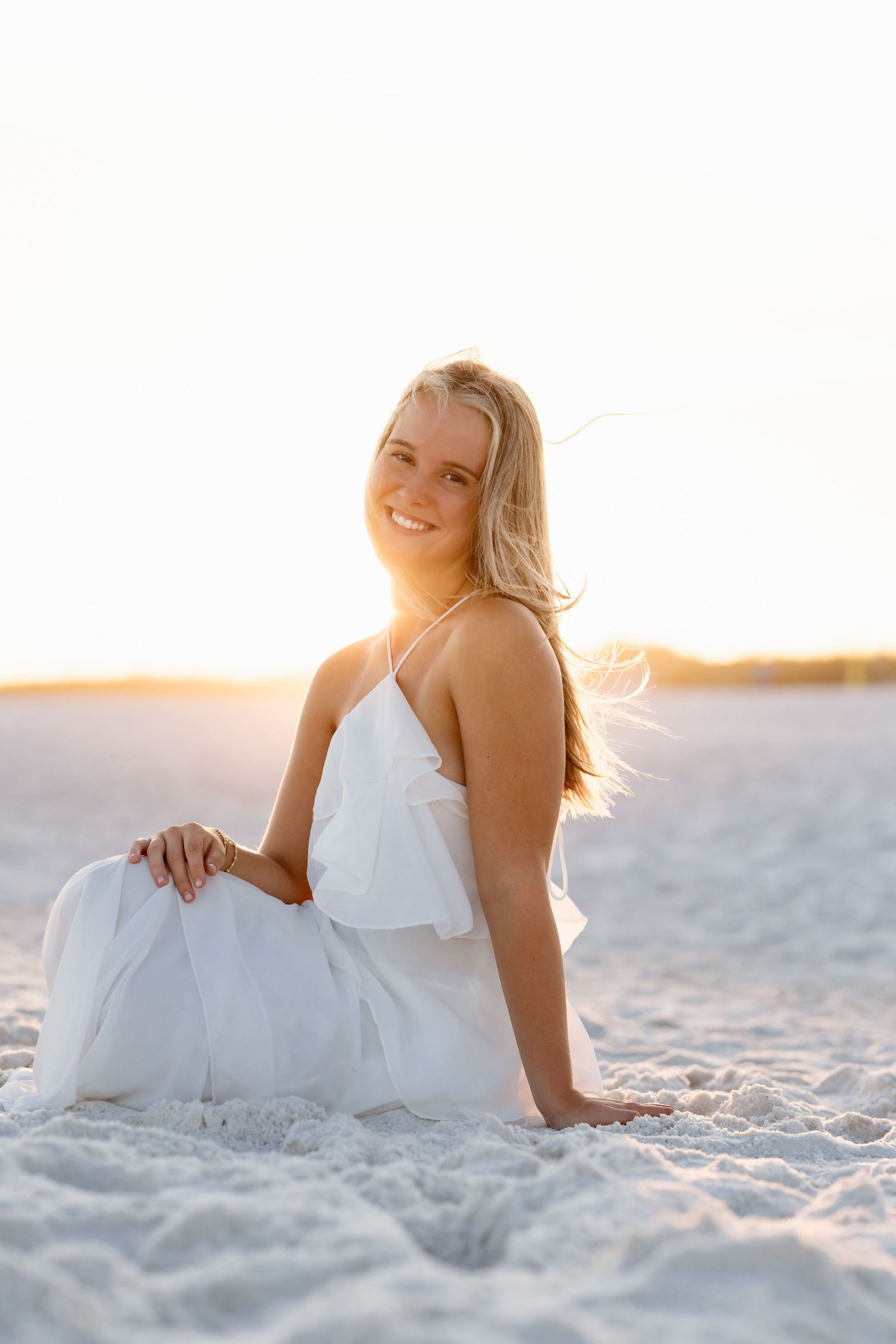 Smiling blonde woman in white ruffled halter dress sitting on white sand beach at golden sunset.