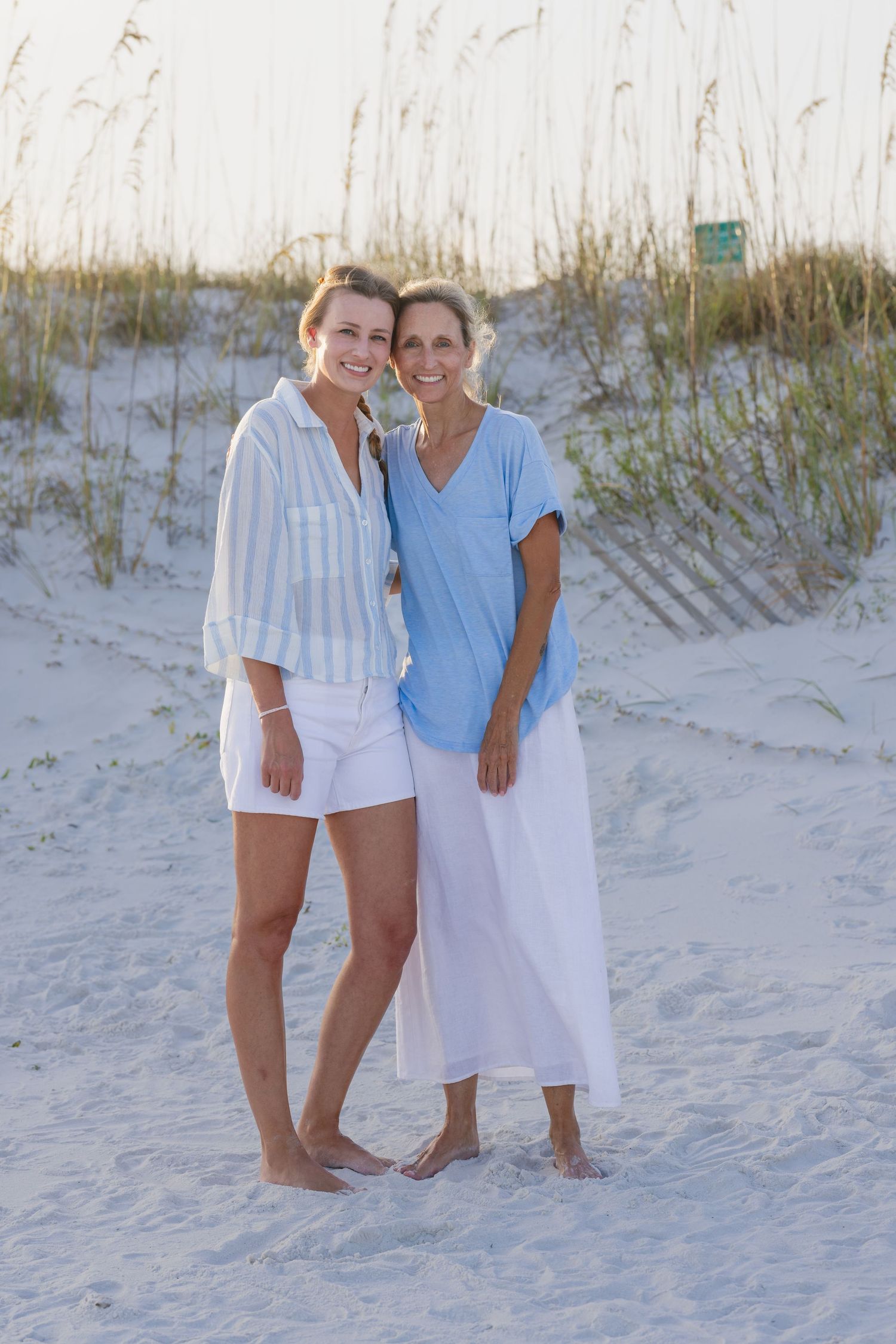 Two women smile together on a sandy beach with sea oats, wearing coordinated blue and white outfits.