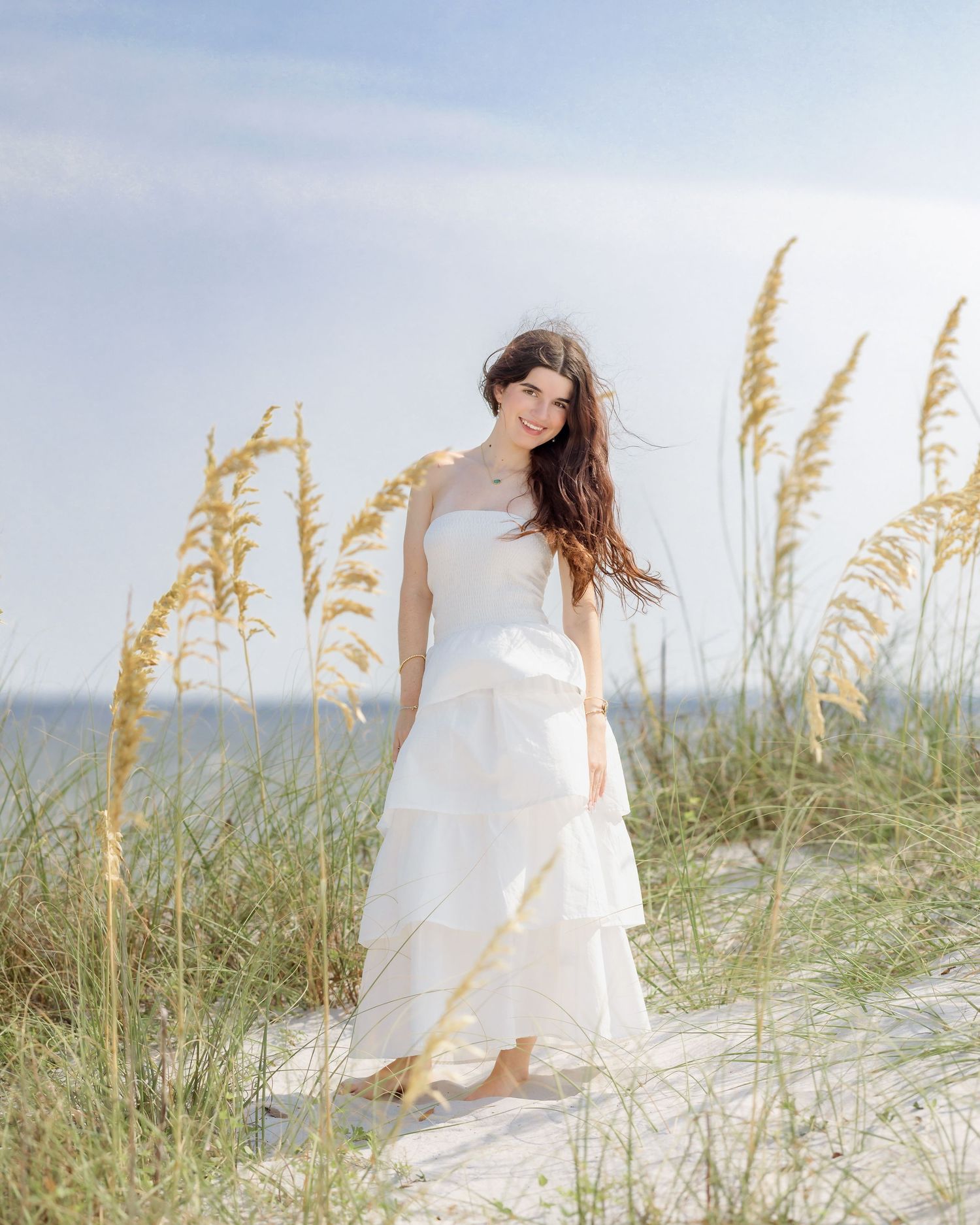 Young woman in white tiered dress standing among tall golden sea oats on beach dunes.