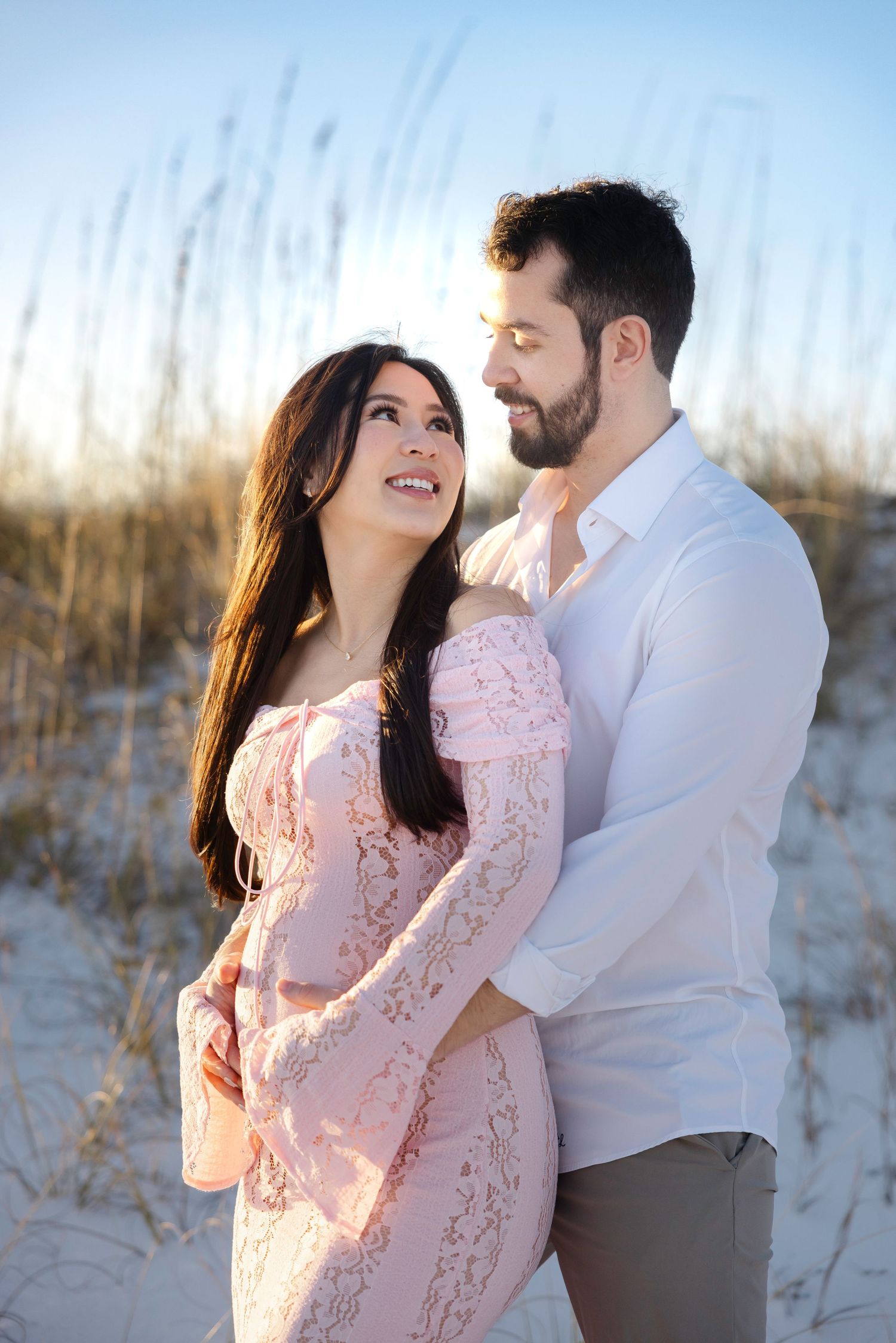 A couple embraces on the beach during golden hour with coastal dunes and beach grass in soft lighting.