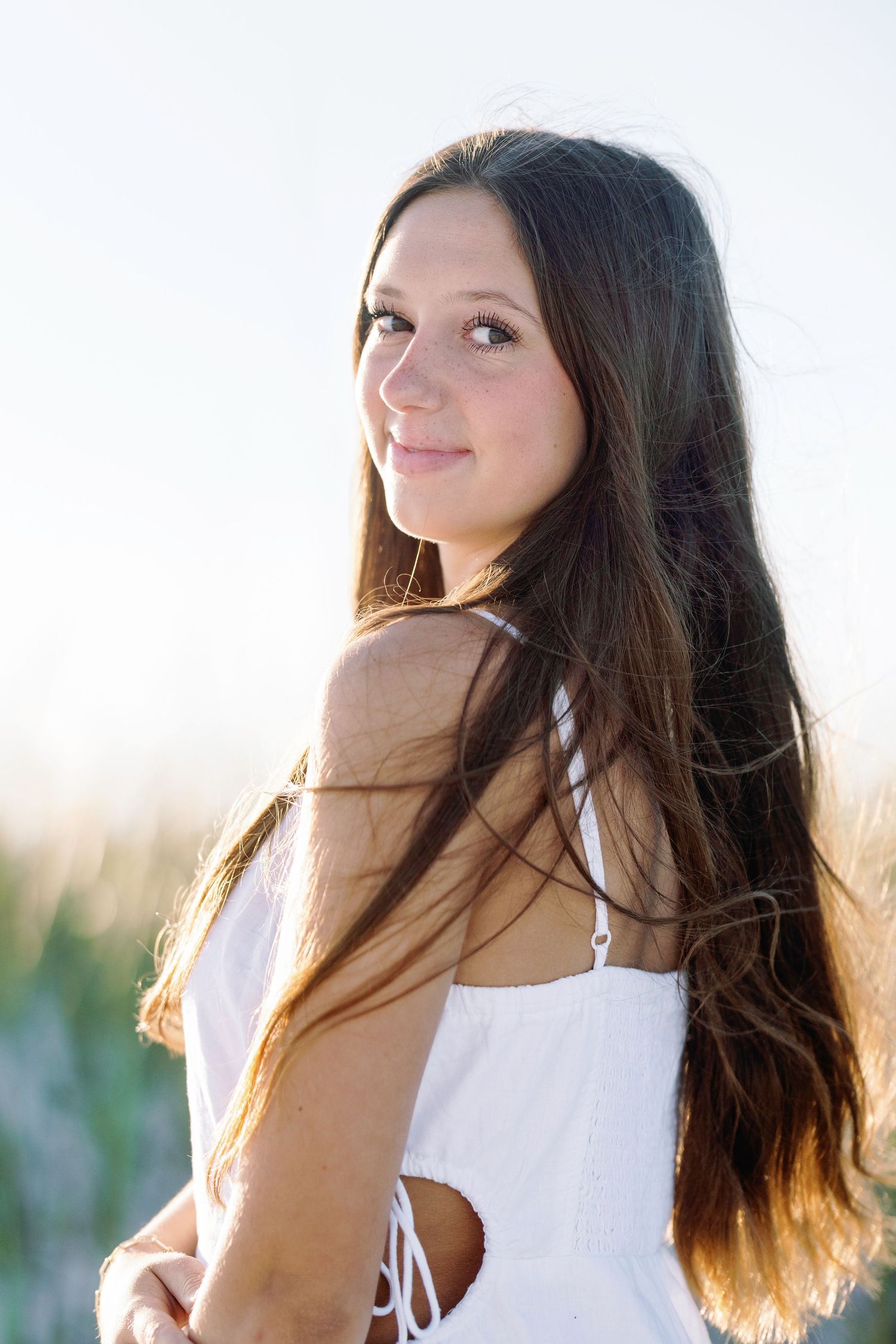 Woman with long dark hair poses in white dress against sunset backdrop in dreamy outdoor portrait session.