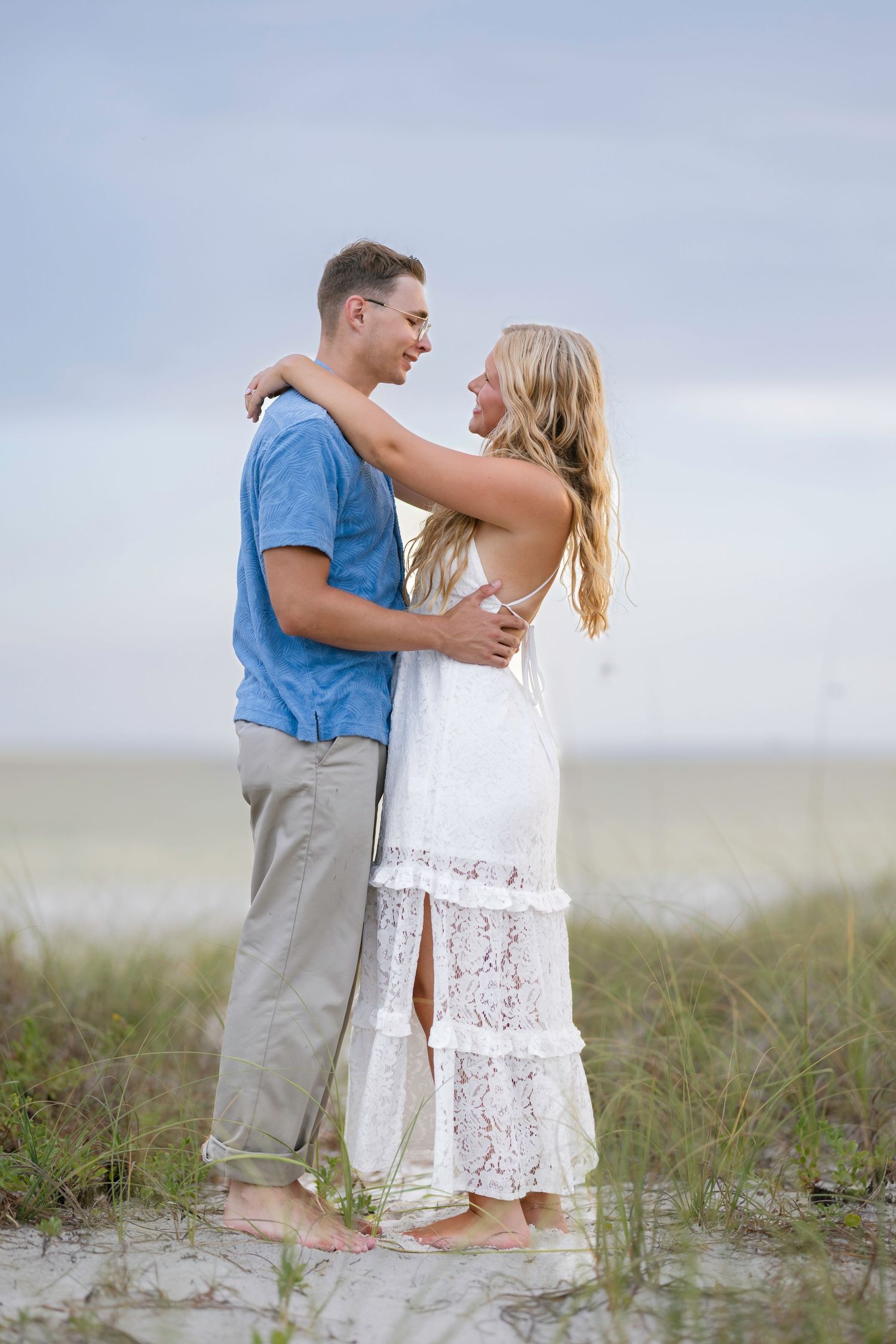 Romantic couple embracing barefoot on sandy beach dunes, woman in white lace dress and man in blue shirt.
