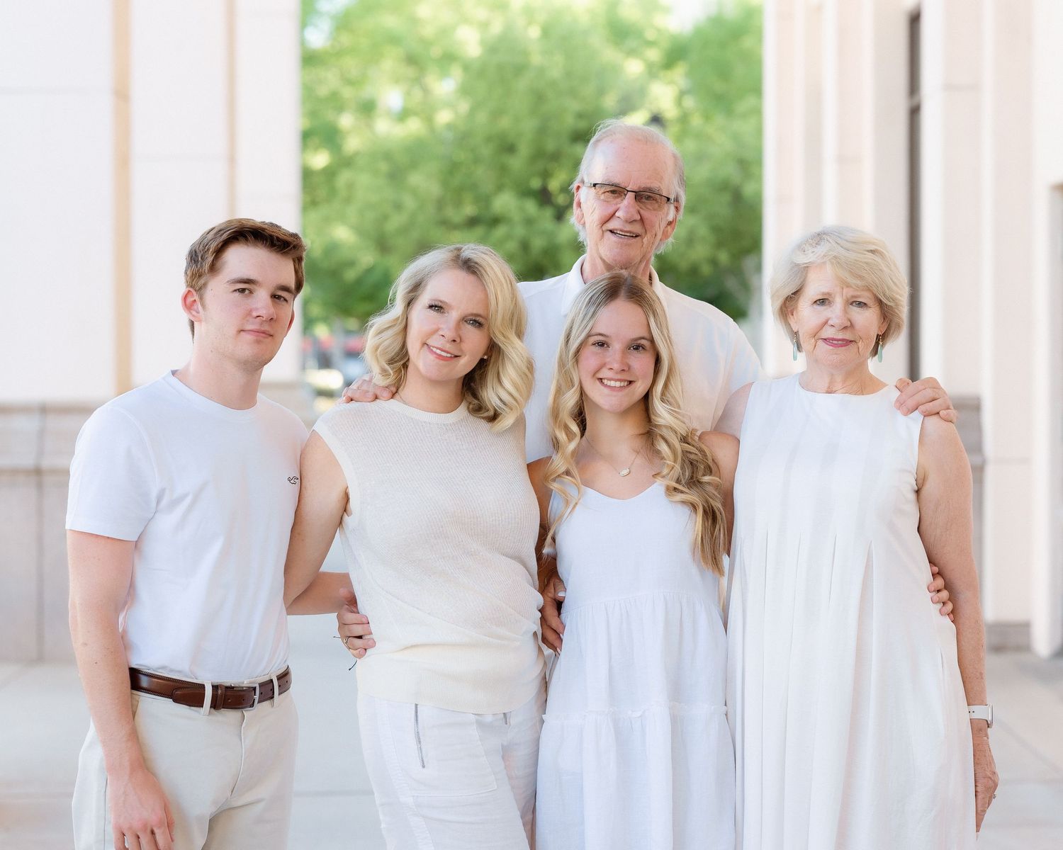 Family of five smiling together in matching white outfits outdoors near columns.