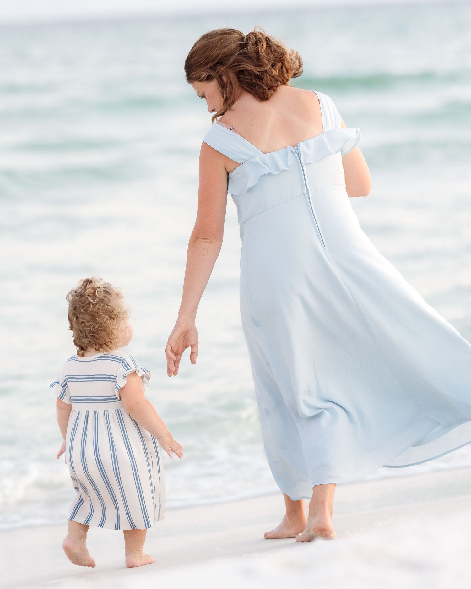 A mother in a light blue off-shoulder gown walks barefoot on a sandy beach with her toddler in a striped dress.