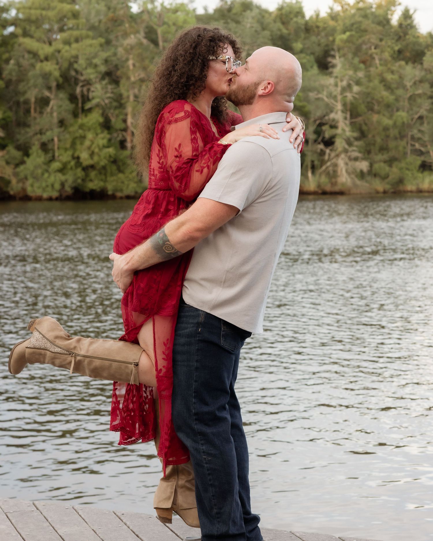 Couple sharing a romantic kiss on a dock by a lake, man lifting woman in red lace dress.