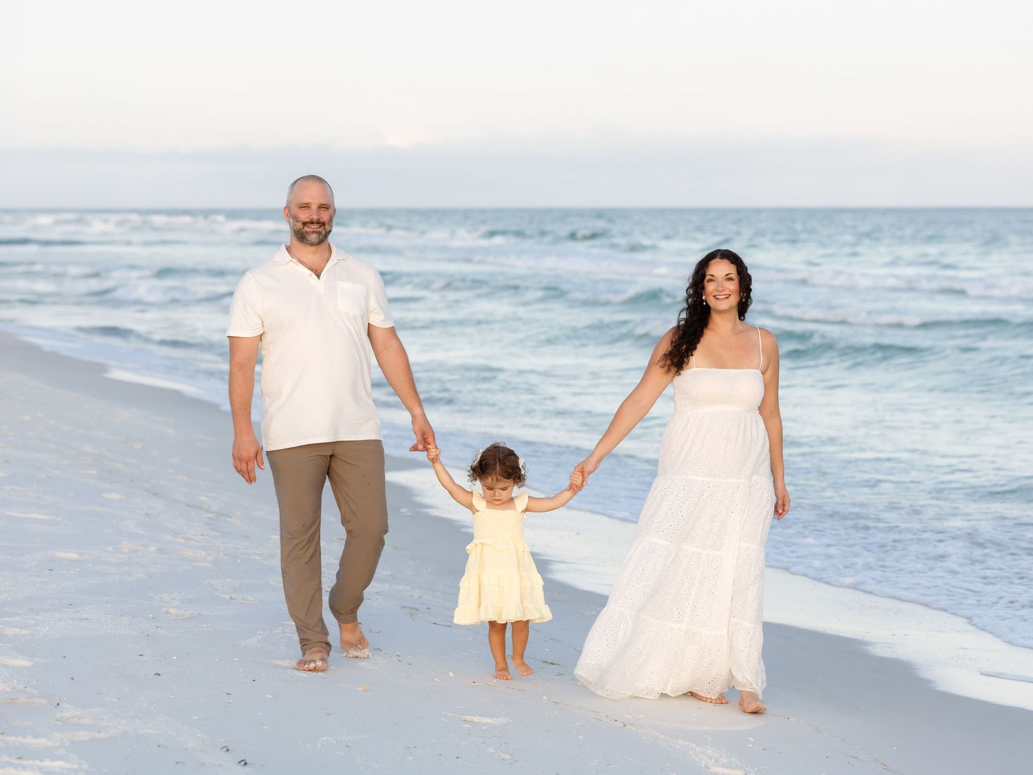 Family of three holding hands walking on a beach, parents in white outfits with toddler in yellow dress.