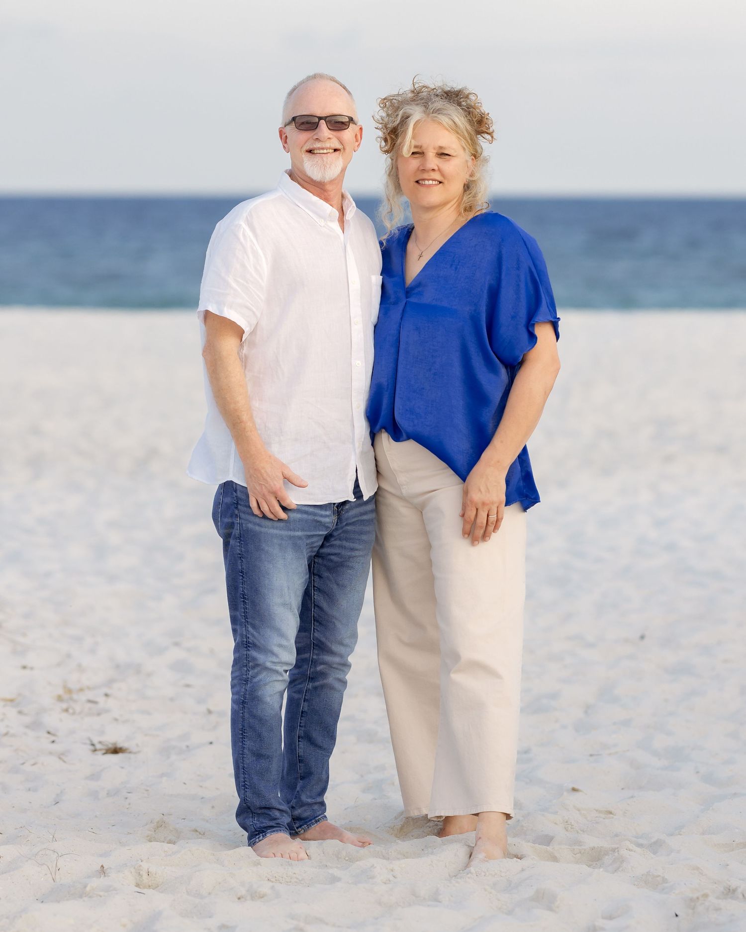 Smiling couple in casual attire posing together on a white sand beach with ocean backdrop.