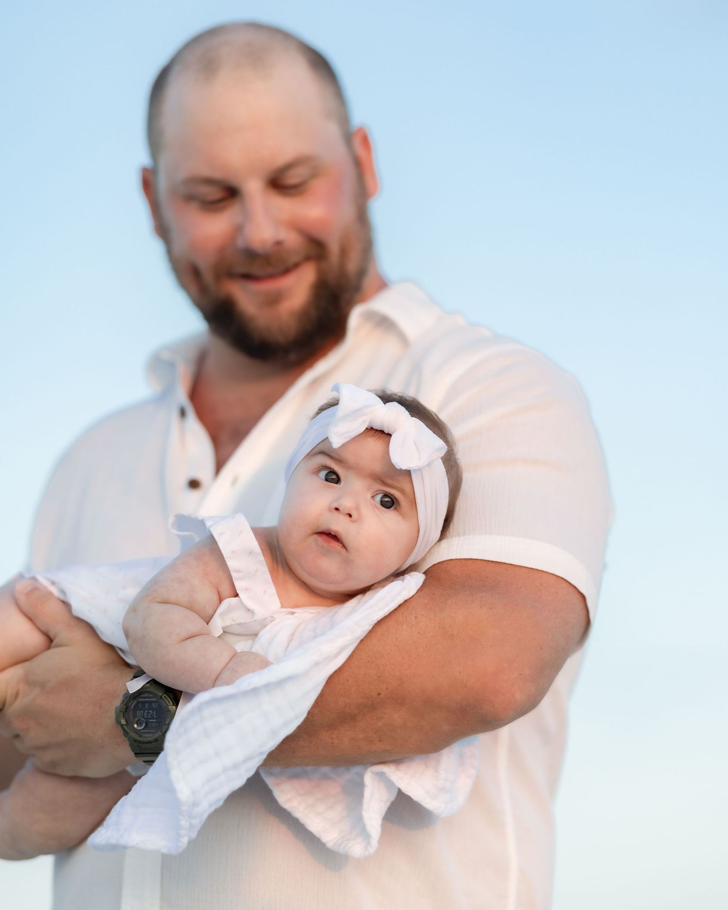 Bearded man in white shirt tenderly holding alert baby with white bow headband outdoors.