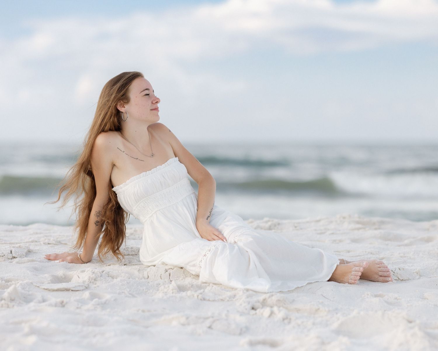 A young woman in a white strapless dress sits on white sand beach with ocean waves behind her.
