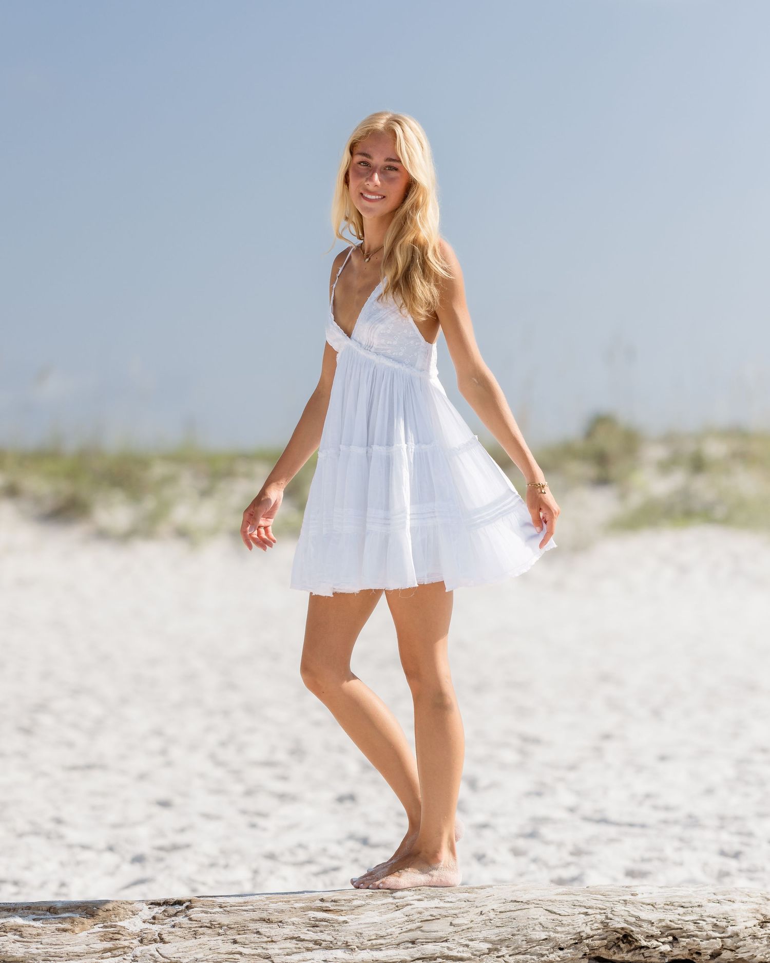 Smiling blonde woman in white mini dress walking barefoot on sandy beach on sunny day.