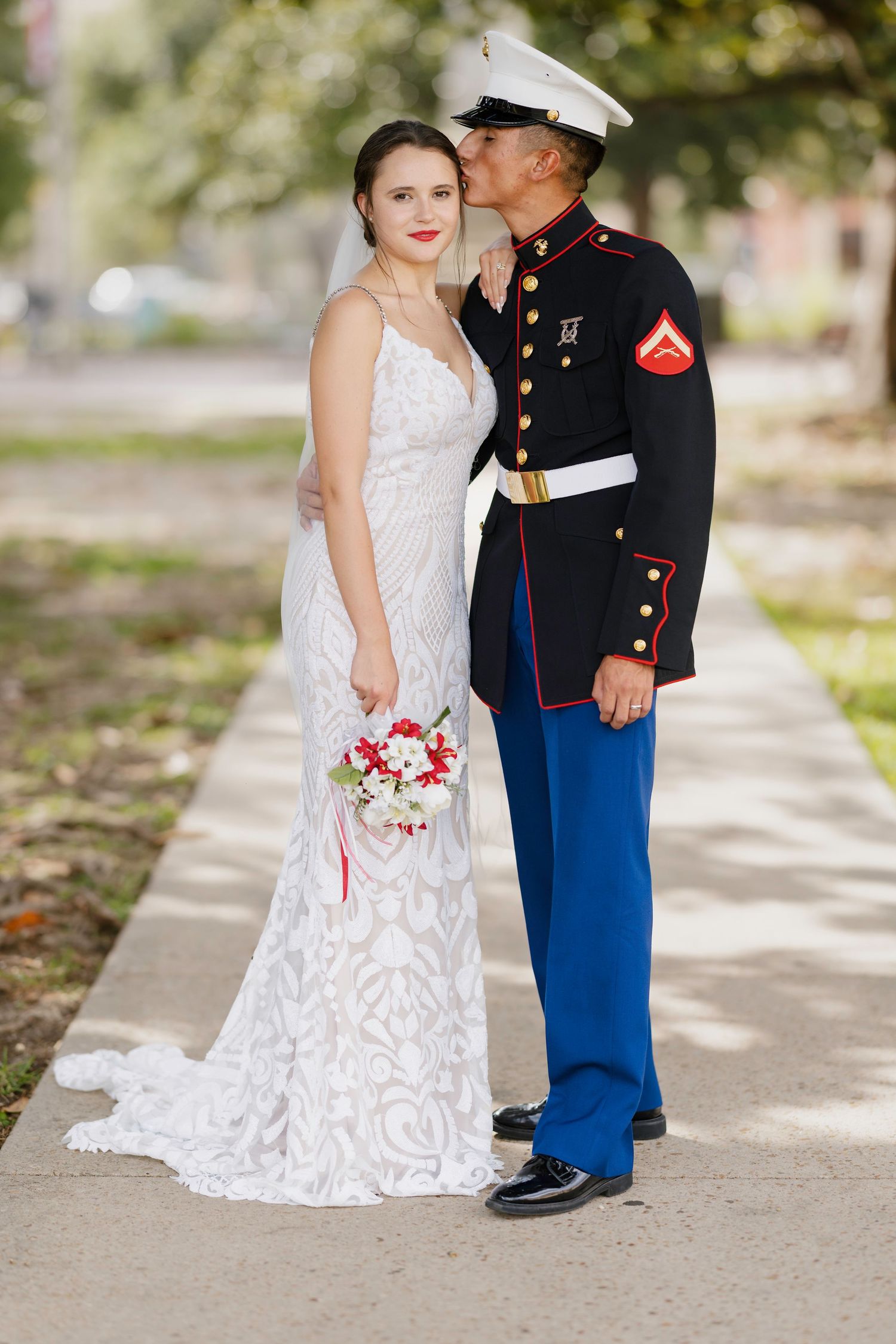 Marine in dress blues kisses bride in lace gown on cheek while she holds red and white bouquet outdoors.