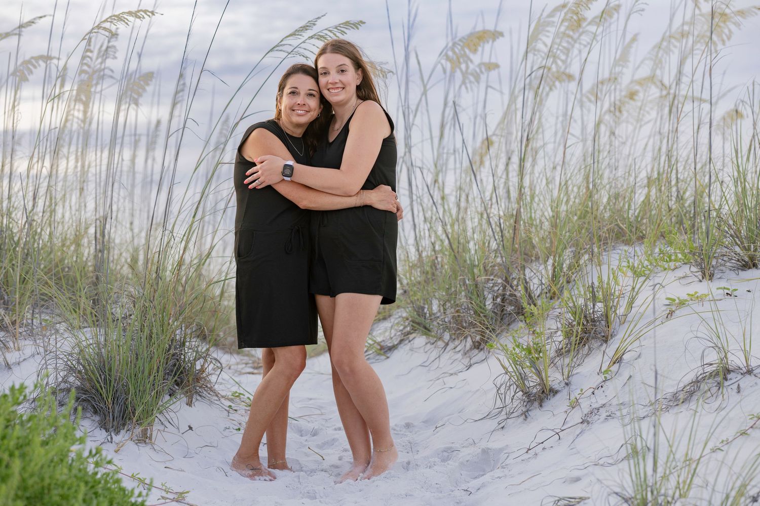 Two young women embrace and smile while standing on white sand dunes with sea oats behind them.